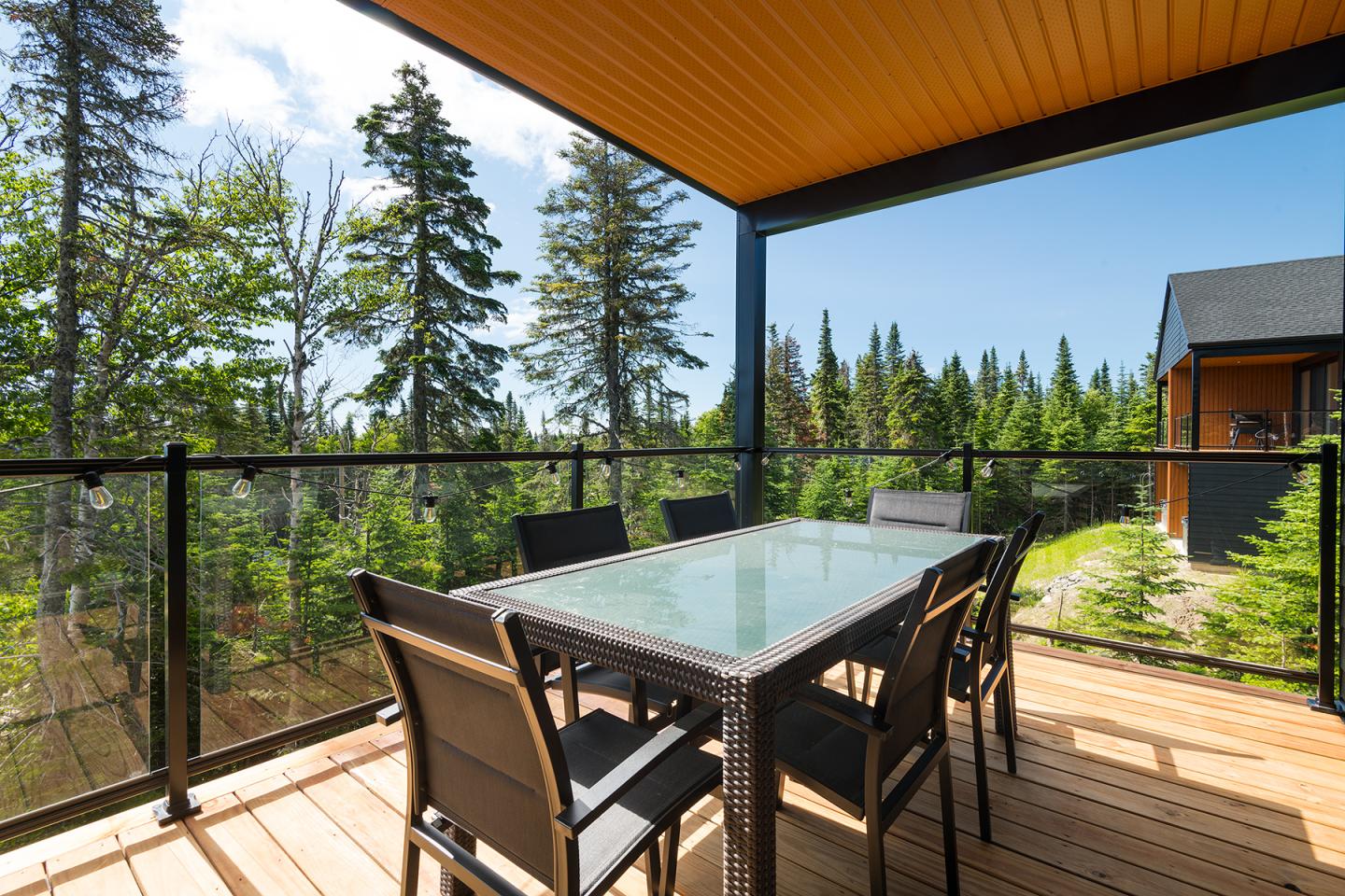 Table en verre et chaises sur une terrasse en bois, entourée d'arbres et sous un ciel bleu.