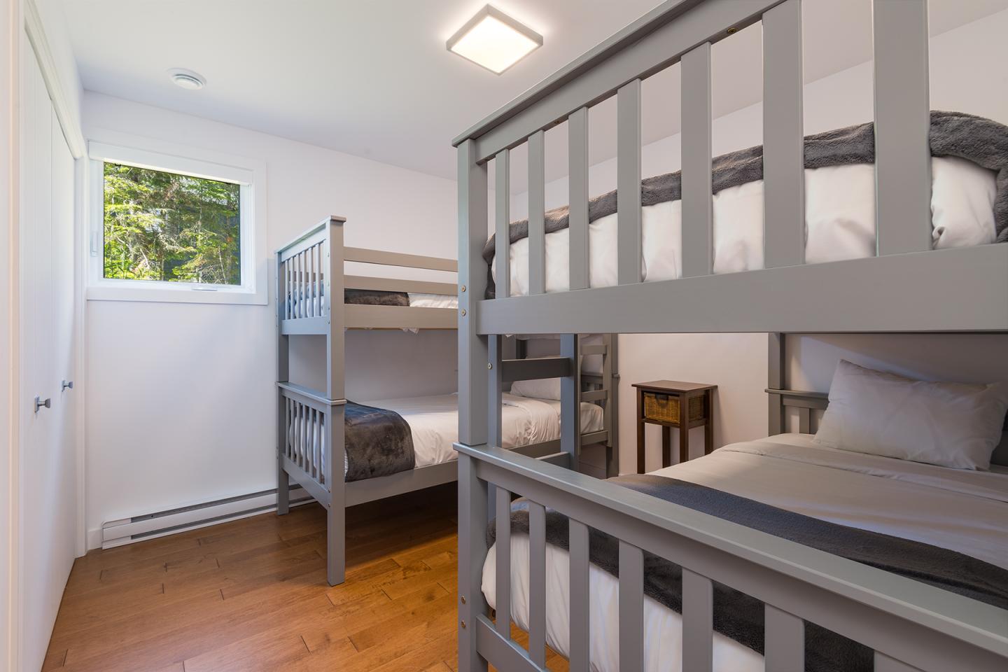 Chambre lumineuse avec lits superposés gris et parquet en bois. Fenêtre à gauche.