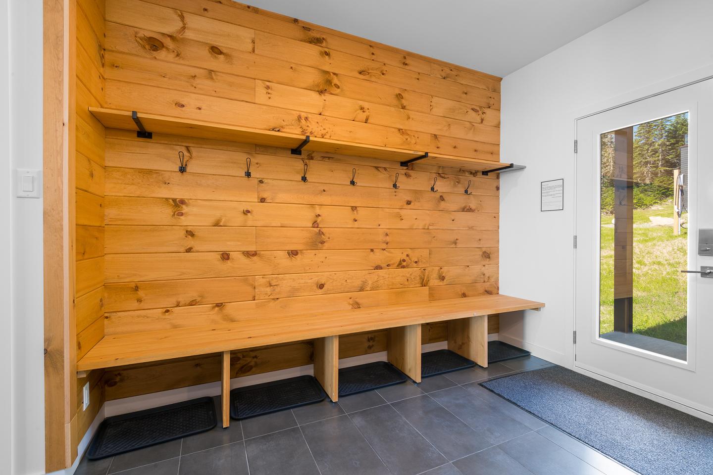 Vestiaire en bois avec bancs et crochets, lumière naturelle par une porte vitrée.