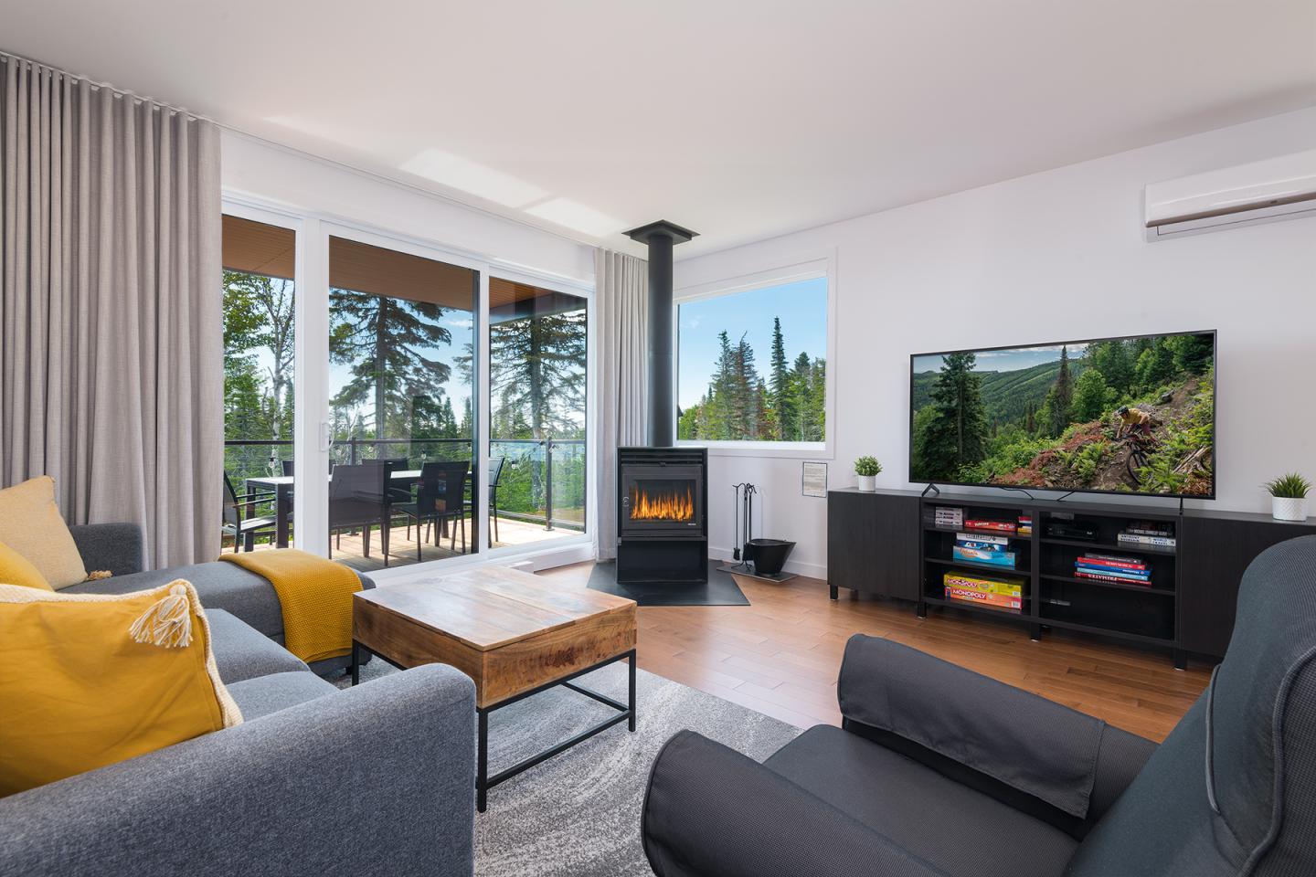 Salon moderne avec canapé gris, cheminée, vue sur terrasse et forêt par la baie vitrée.