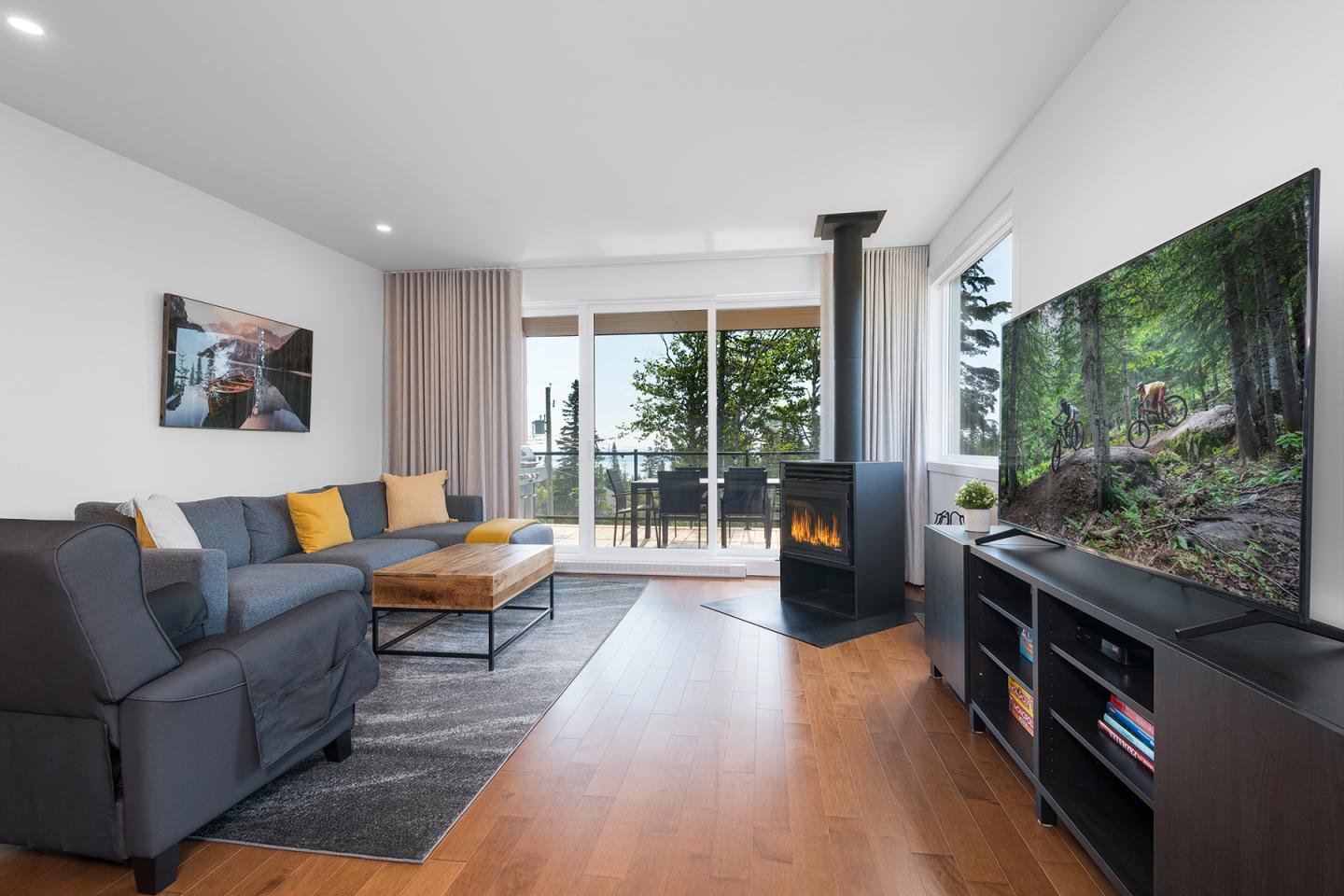 Salon moderne avec canapé gris, poêle noir, et grande TV. Vue sur balcon et arbres.