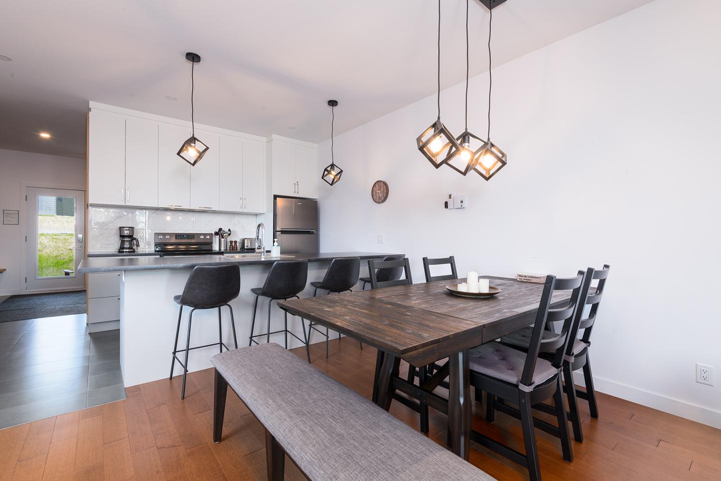 Cuisine moderne avec table en bois, chaises noires et éclairage suspendu.