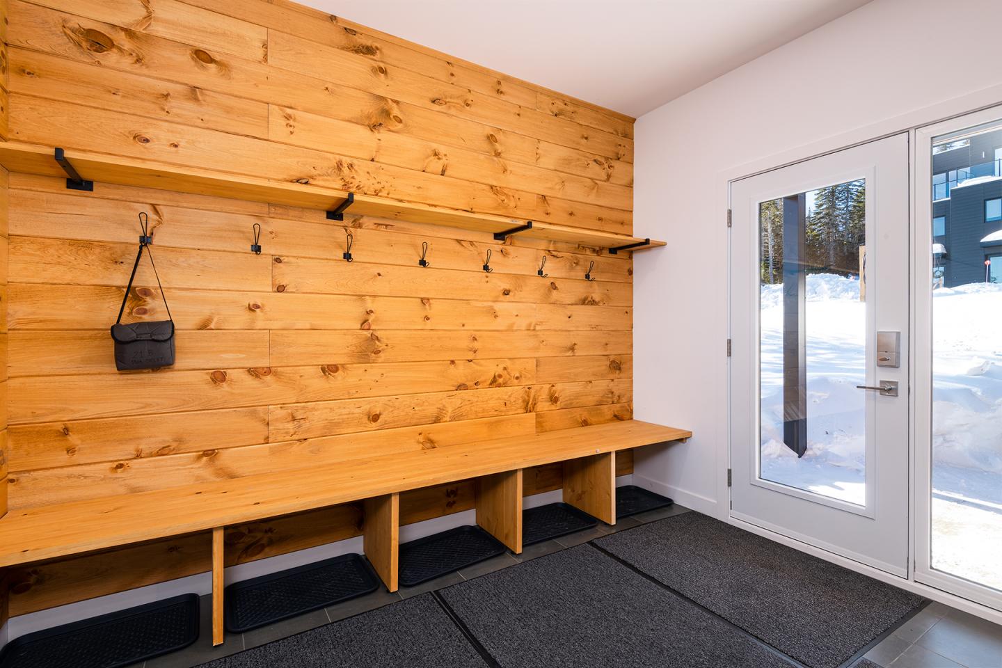 Intérieur en bois avec banc, crochets et sac suspendu, vue sur la neige par la porte vitrée.