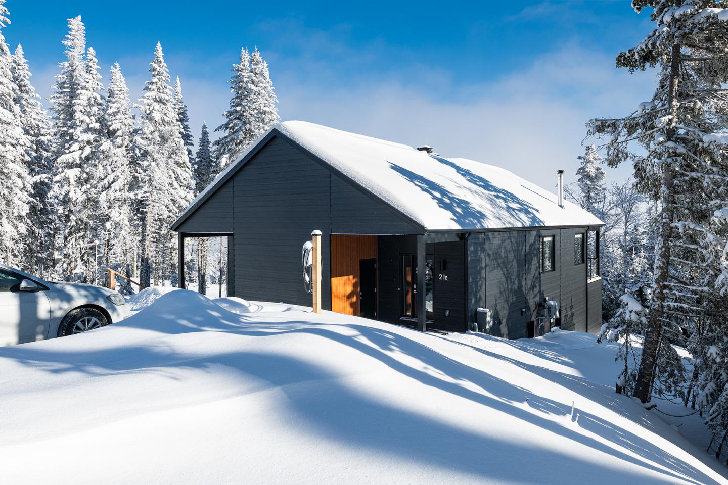 Cabane noire dans la neige, entourée d'arbres enneigés sous un ciel bleu.