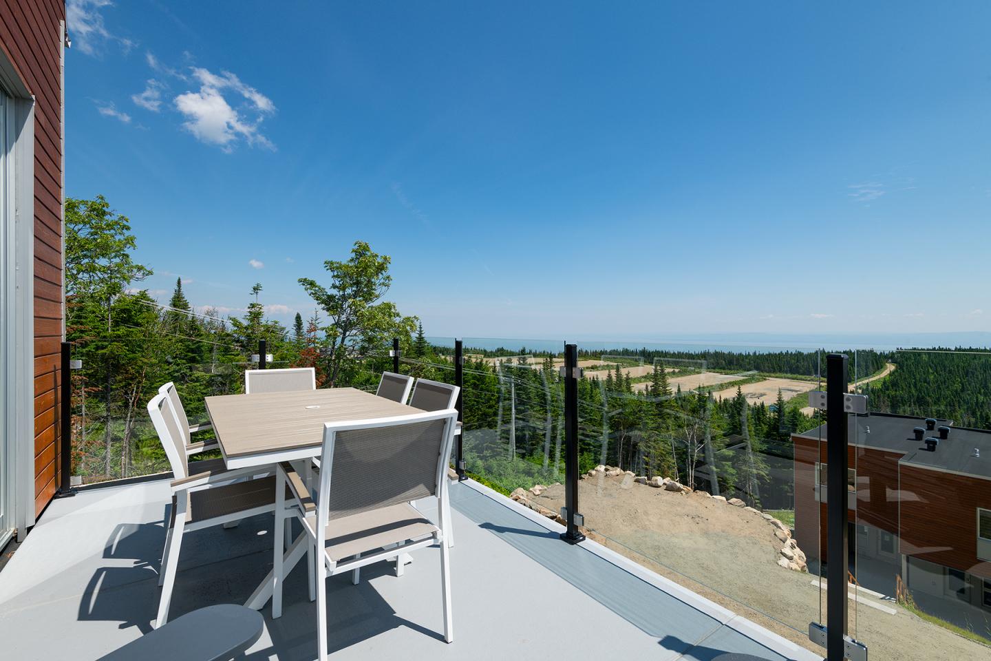 Terrasse avec table et chaises, vue sur forêt et ciel bleu.