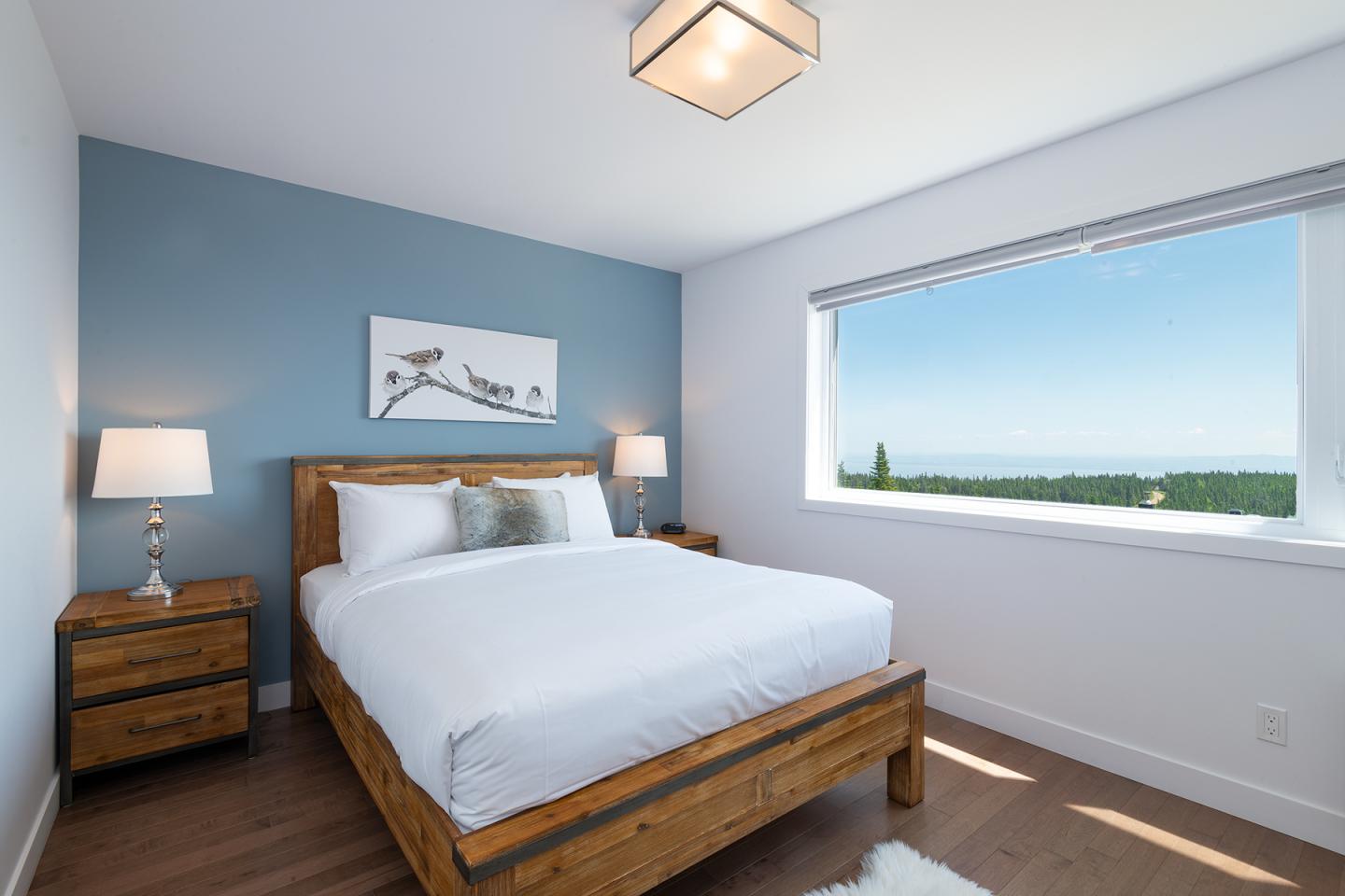 Chambre lumineuse, lit en bois, mur bleu, grande fenêtre avec vue sur la forêt.