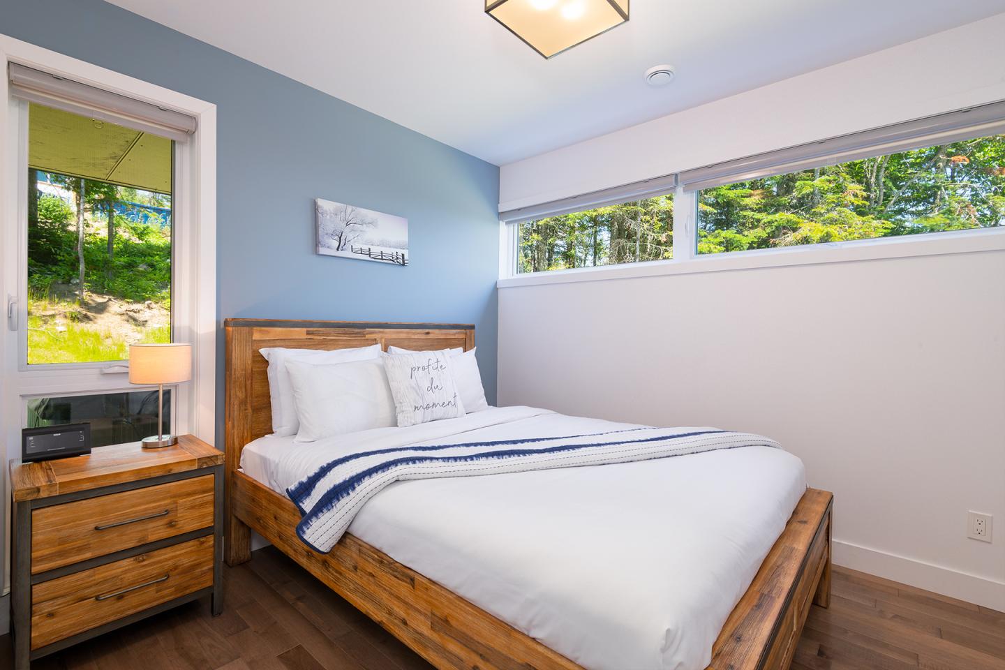 Chambre lumineuse avec lit double en bois, mur bleu, fenêtre donnant sur verdure.