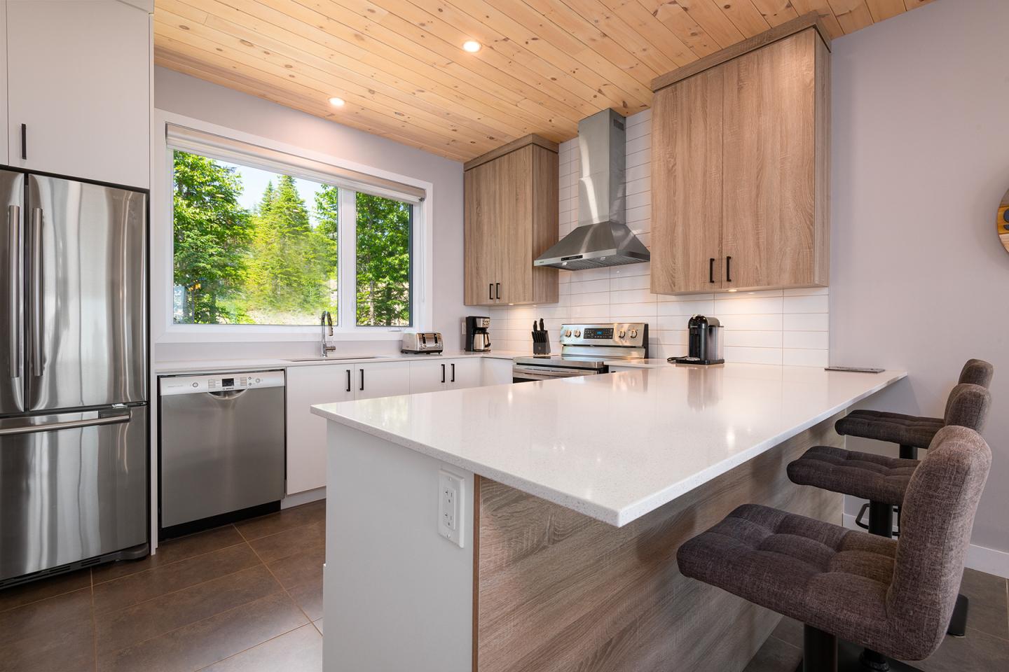 Cuisine moderne avec comptoirs blancs, armoires en bois et grandes fenêtres.