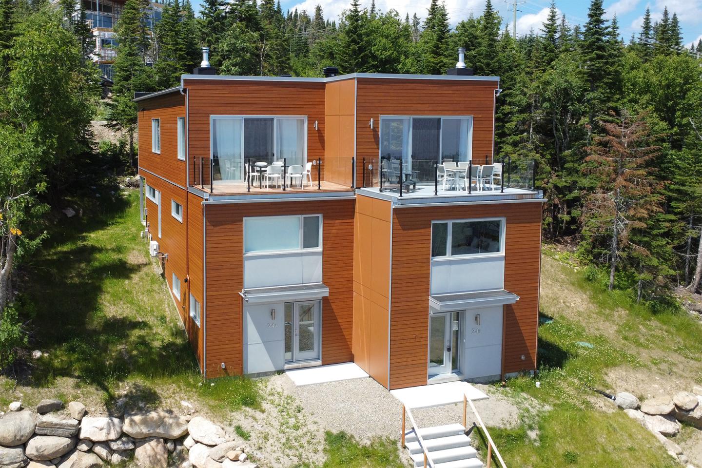 Maison moderne en bois à deux étages, entourée de verdure et de rochers.