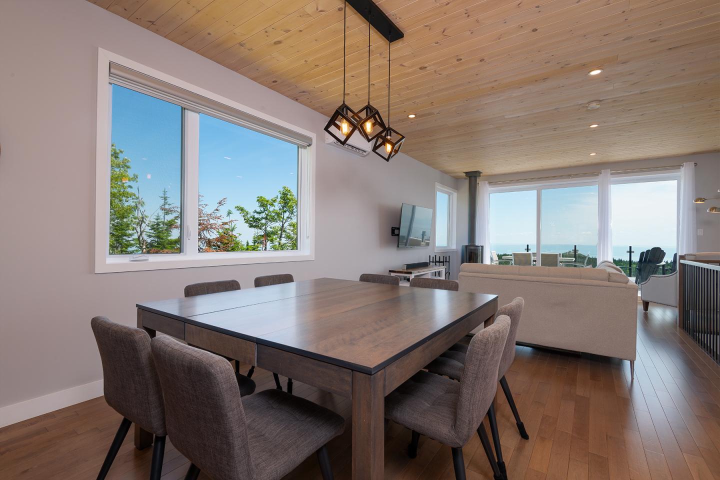 Salle à manger moderne avec table en bois, grandes fenêtres et lumière naturelle.