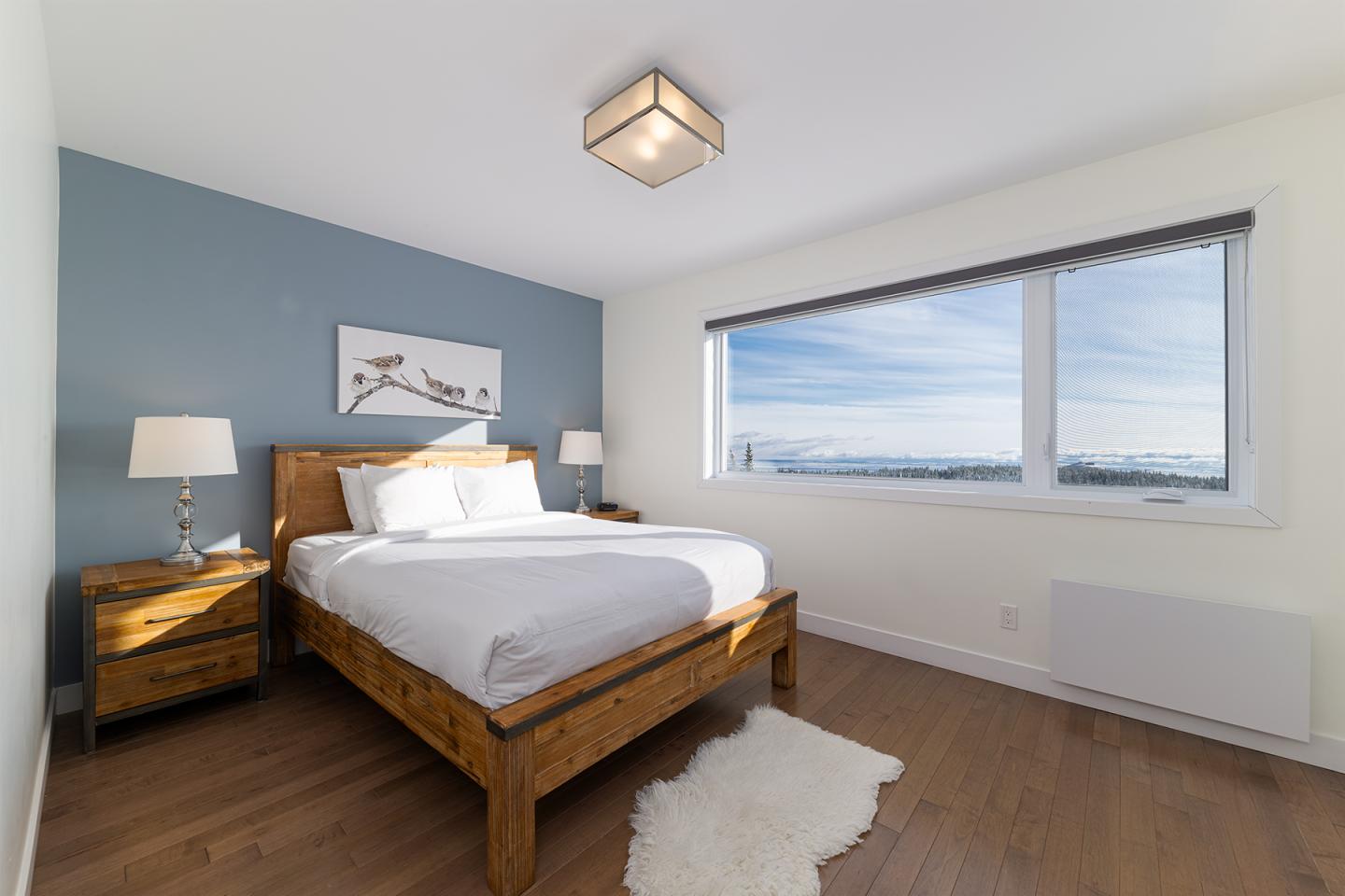 Chambre lumineuse, lit double en bois, mur bleu, grande fenêtre avec vue.