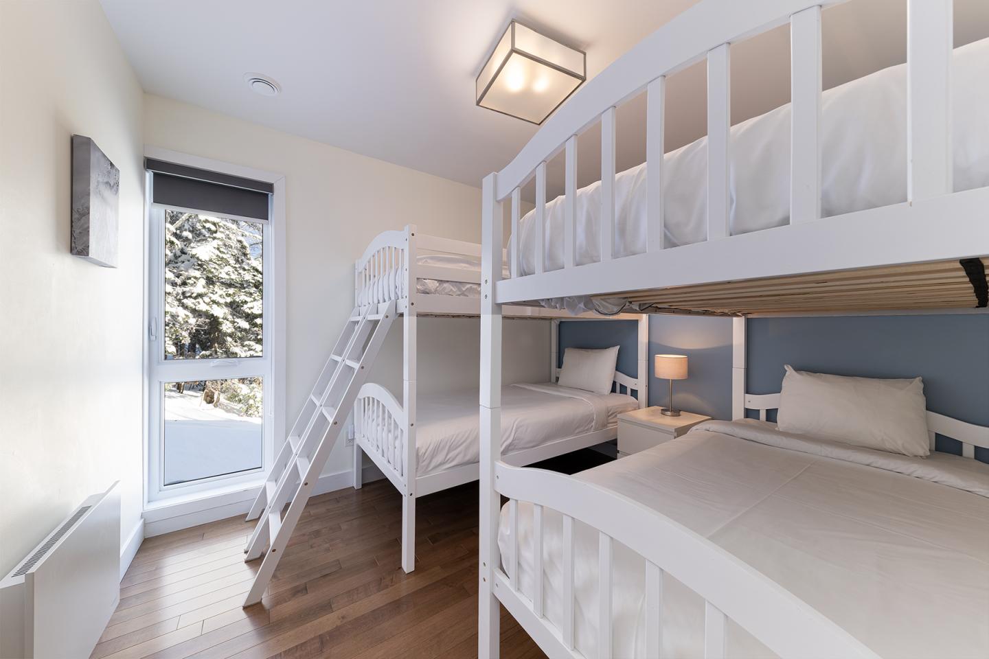 Chambre avec lits superposés blancs, parquet et fenêtre lumineuse.