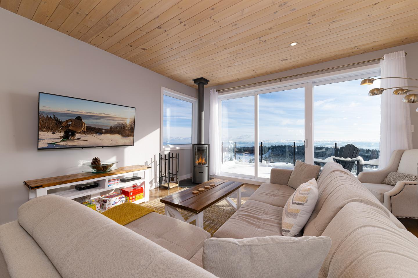 Salon lumineux avec canapé beige, grande fenêtre vue sur la mer, télé et poêle à bois.