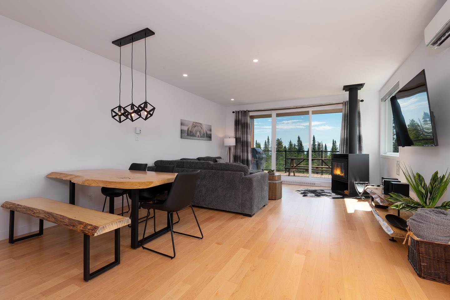 Salon moderne avec cheminée, canapé gris, table en bois et vue sur la forêt.