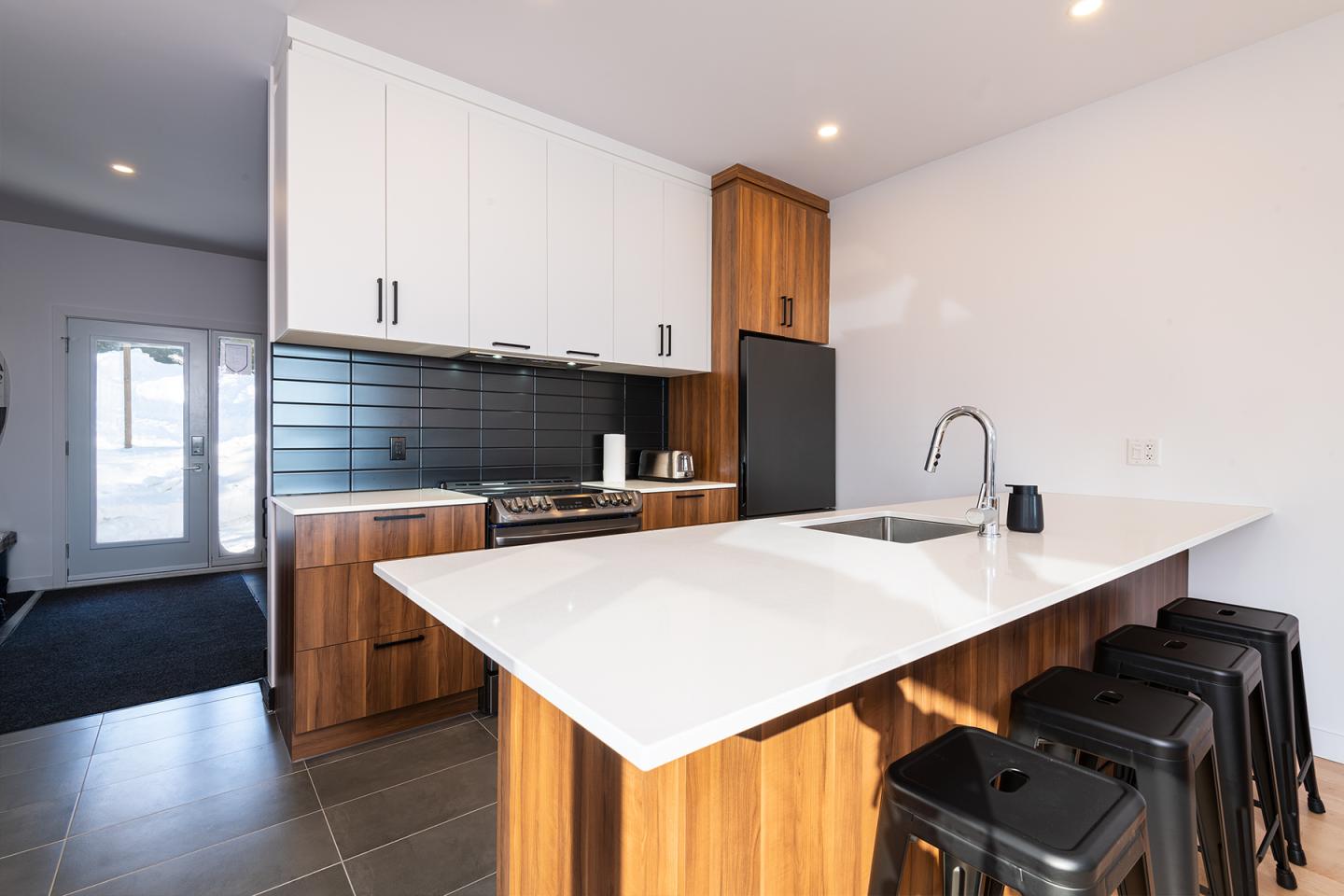 Cuisine moderne avec îlot, armoires bois, mur blanc, tabourets noirs.