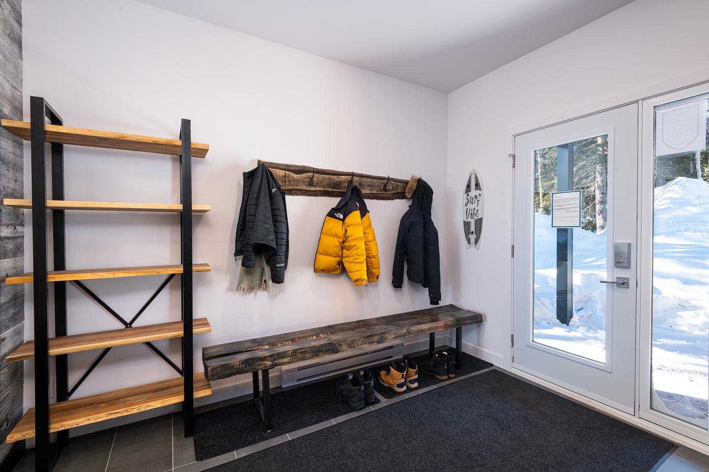 Vestibule lumineux avec étagère en bois, manteaux suspendus et porte vitrée enneigée.
