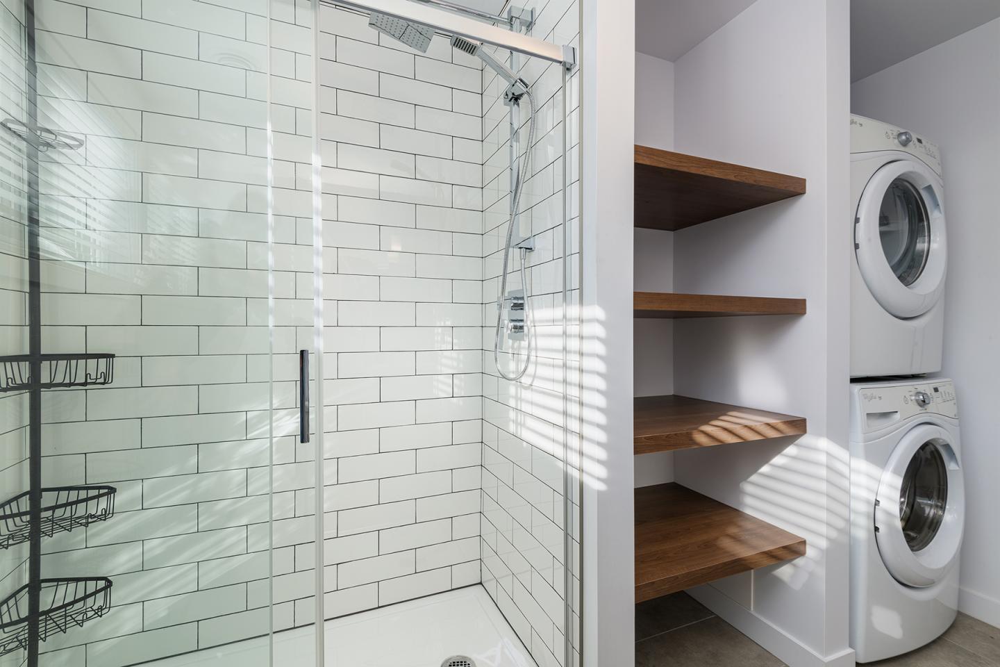 Douche vitrée et lave-linge superposés, étagères en bois.