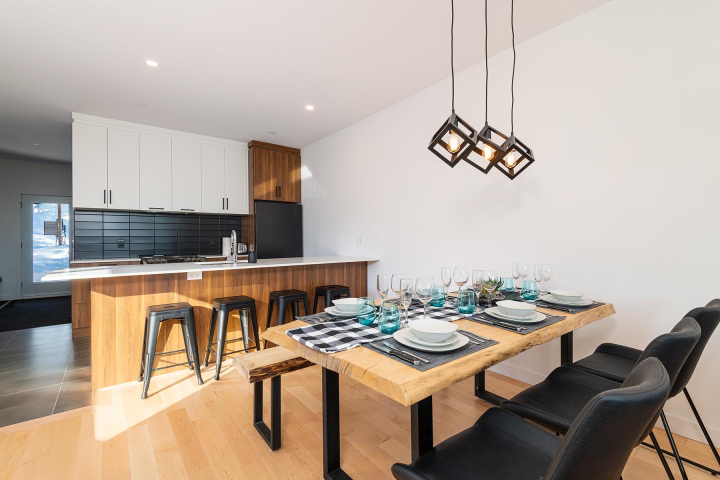 Salle à manger moderne avec table dressée, cuisine ouverte et éclairage suspendu.
