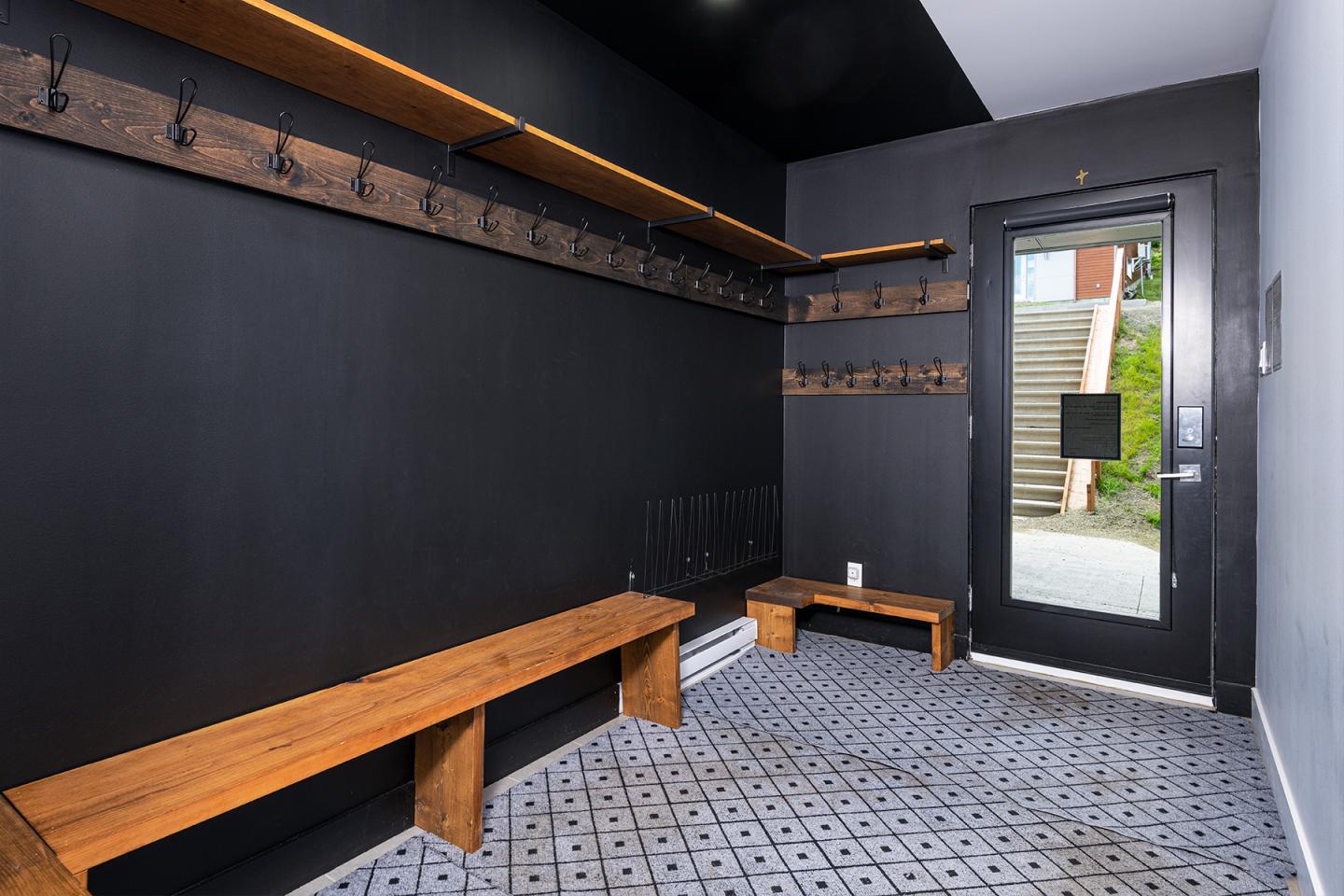 Vestiaire noir avec bancs en bois et crochets, porte menant à l'extérieur.