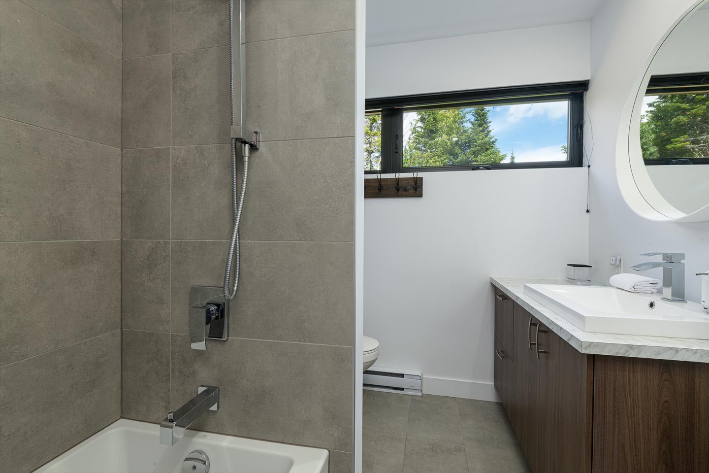 Salle de bain moderne avec douche, lavabo et fenêtre offrant vue sur des arbres.