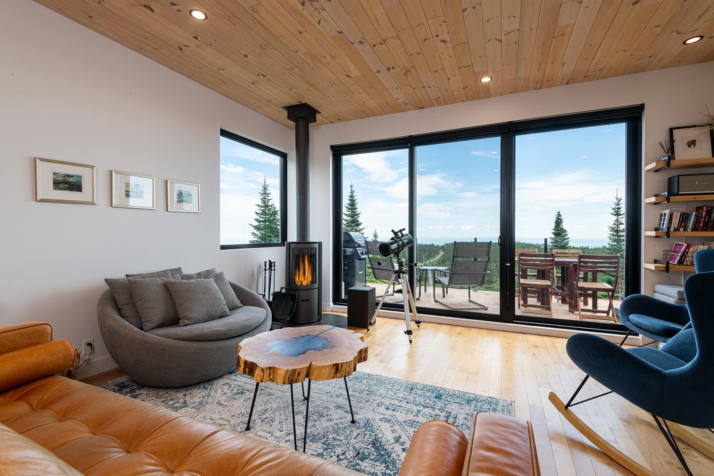 Salon moderne avec cheminée, grandes fenêtres, canapé gris, et vue sur un paysage verdoyant.