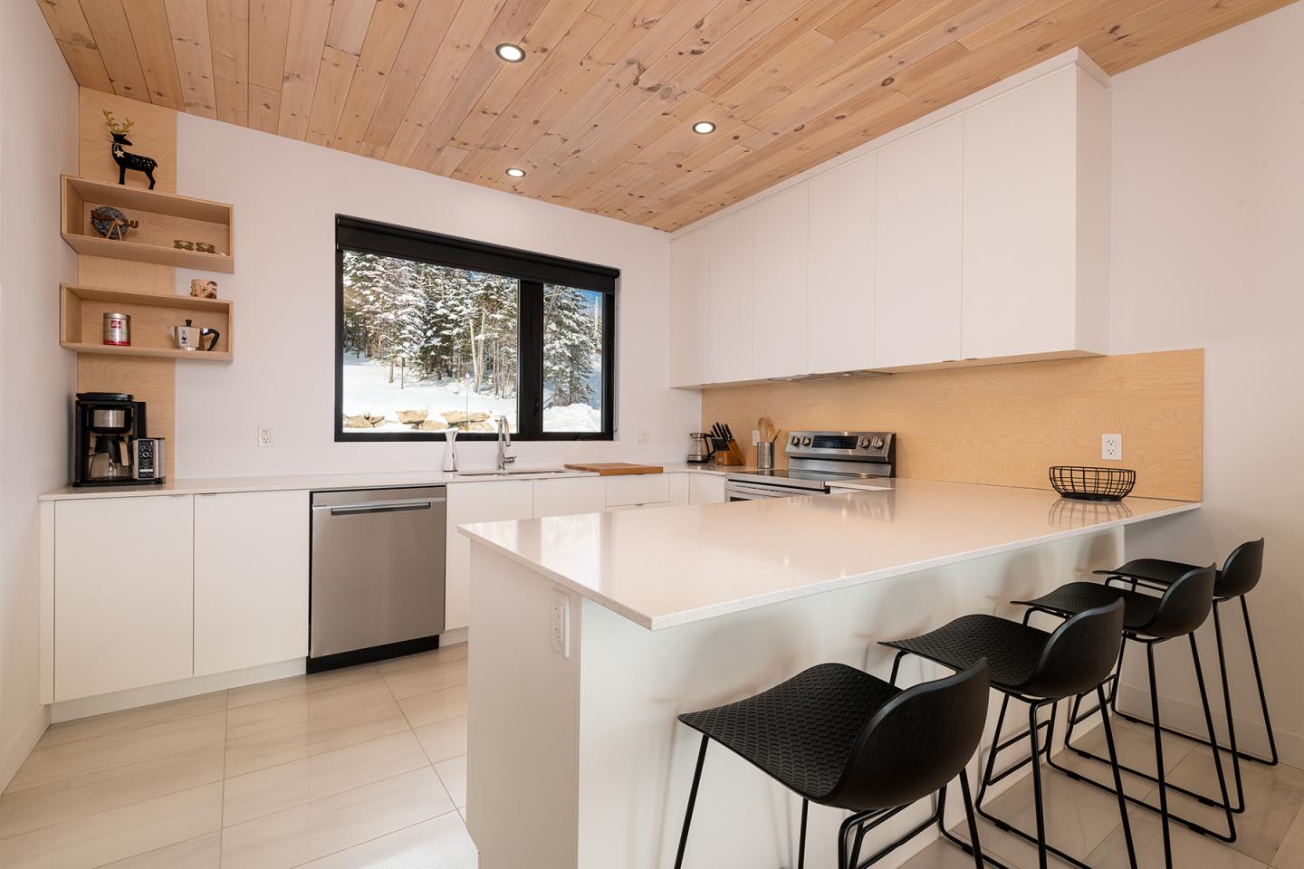 Cuisine moderne blanche avec comptoir-bar, chaises noires, fenêtre donnant sur un extérieur enneigé.