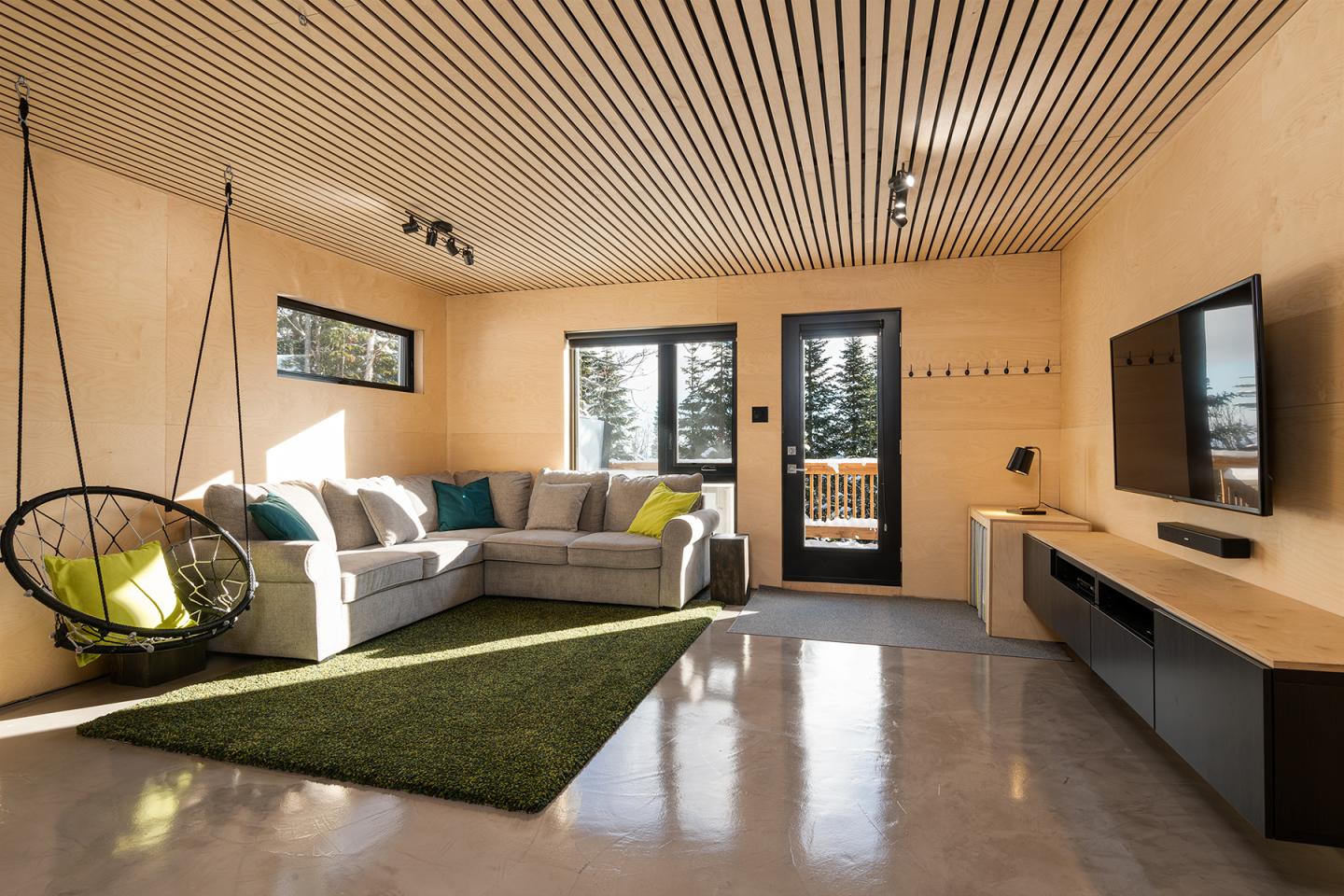 Salon moderne en bois clair avec canapé, TV, et fauteuil suspendu près d'une grande fenêtre.