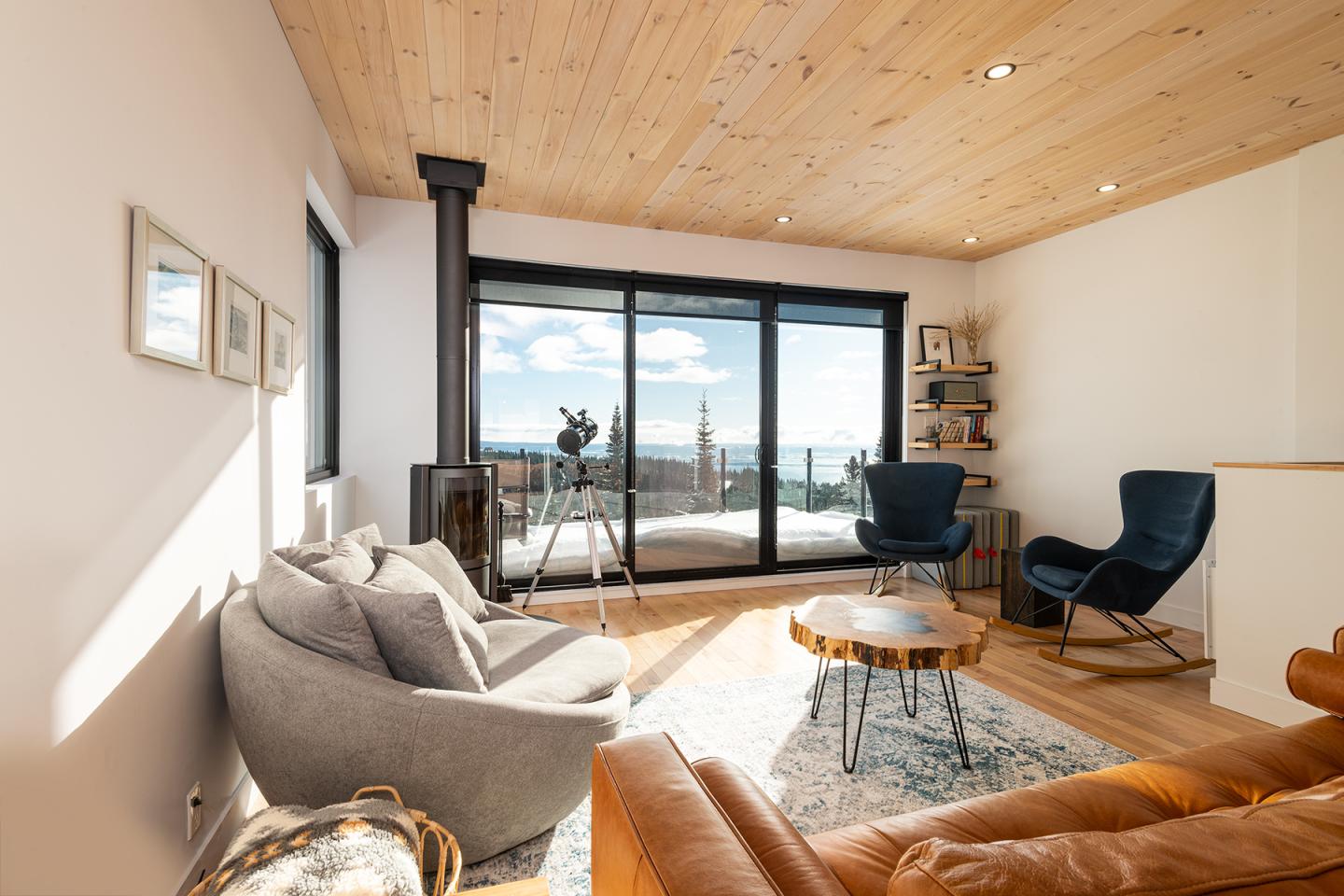 Salon lumineux avec grandes fenêtres, fauteuils bleu et gris, et vue sur la mer.