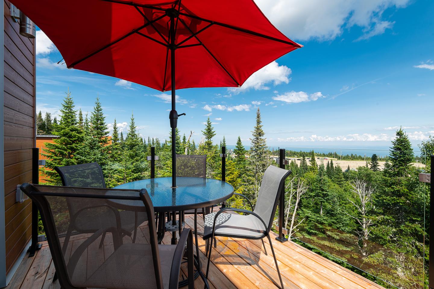 Terrasse en bois avec parasol rouge et vue sur forêt et ciel bleu.
