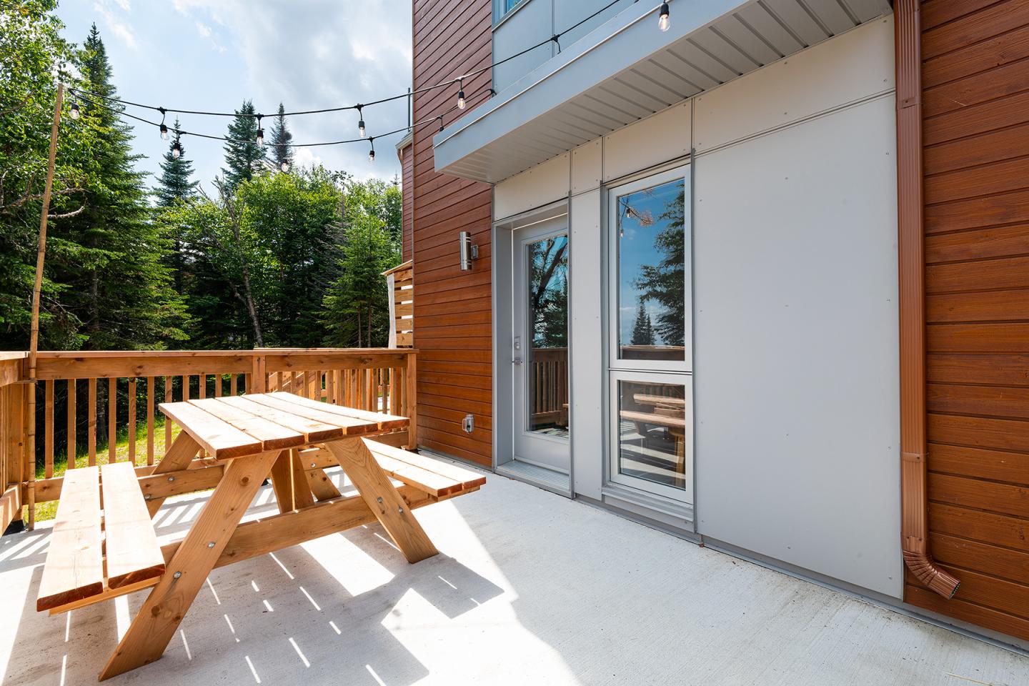 Terrasse en bois avec une table de pique-nique, entourée d'arbres sous un ciel partiellement nuageux.