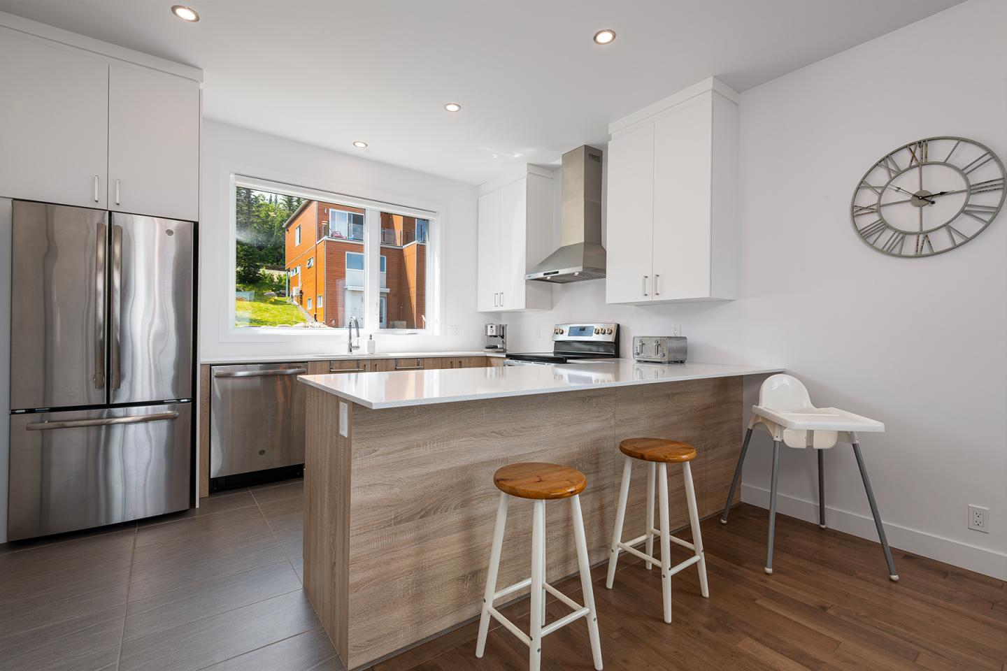 Cuisine moderne avec îlot central, tabourets, et électroménagers en inox. Fenêtre en arrière-plan.
