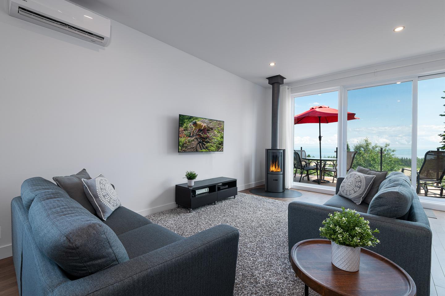 Salon moderne avec canapés gris, cheminée, TV, et vue sur terrasse avec parasol rouge.
