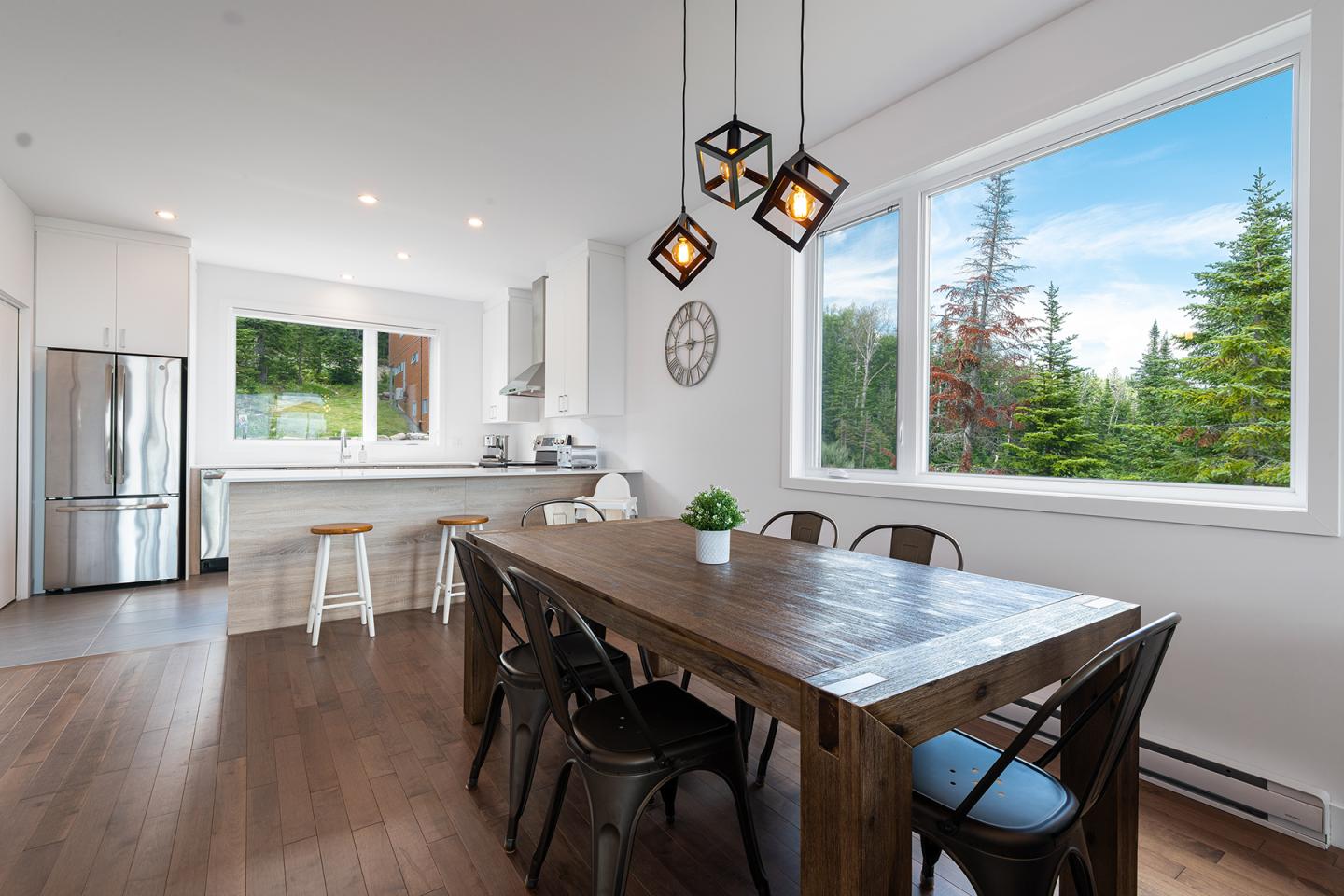 Salle à manger moderne avec table en bois, luminaires suspendus, vue sur forêt.