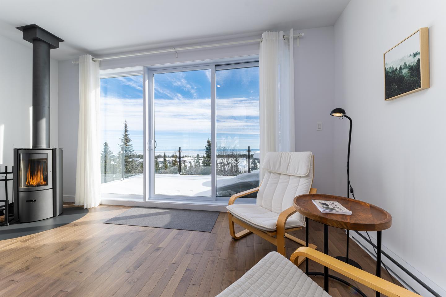 Salon lumineux avec poêle à bois, chaise blanche, vue sur neige.