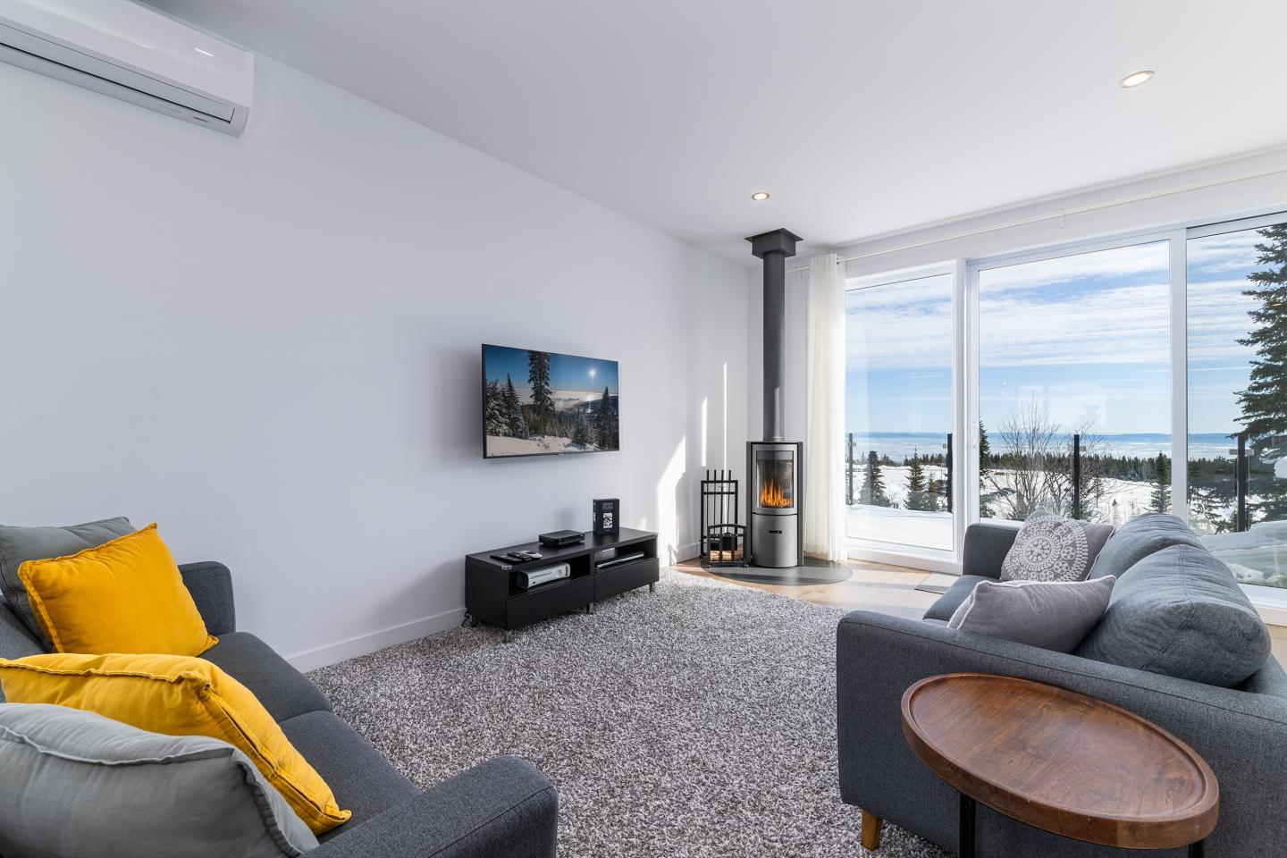 Salon moderne avec vue sur mer, doté de deux canapés gris et un poêle à bois.