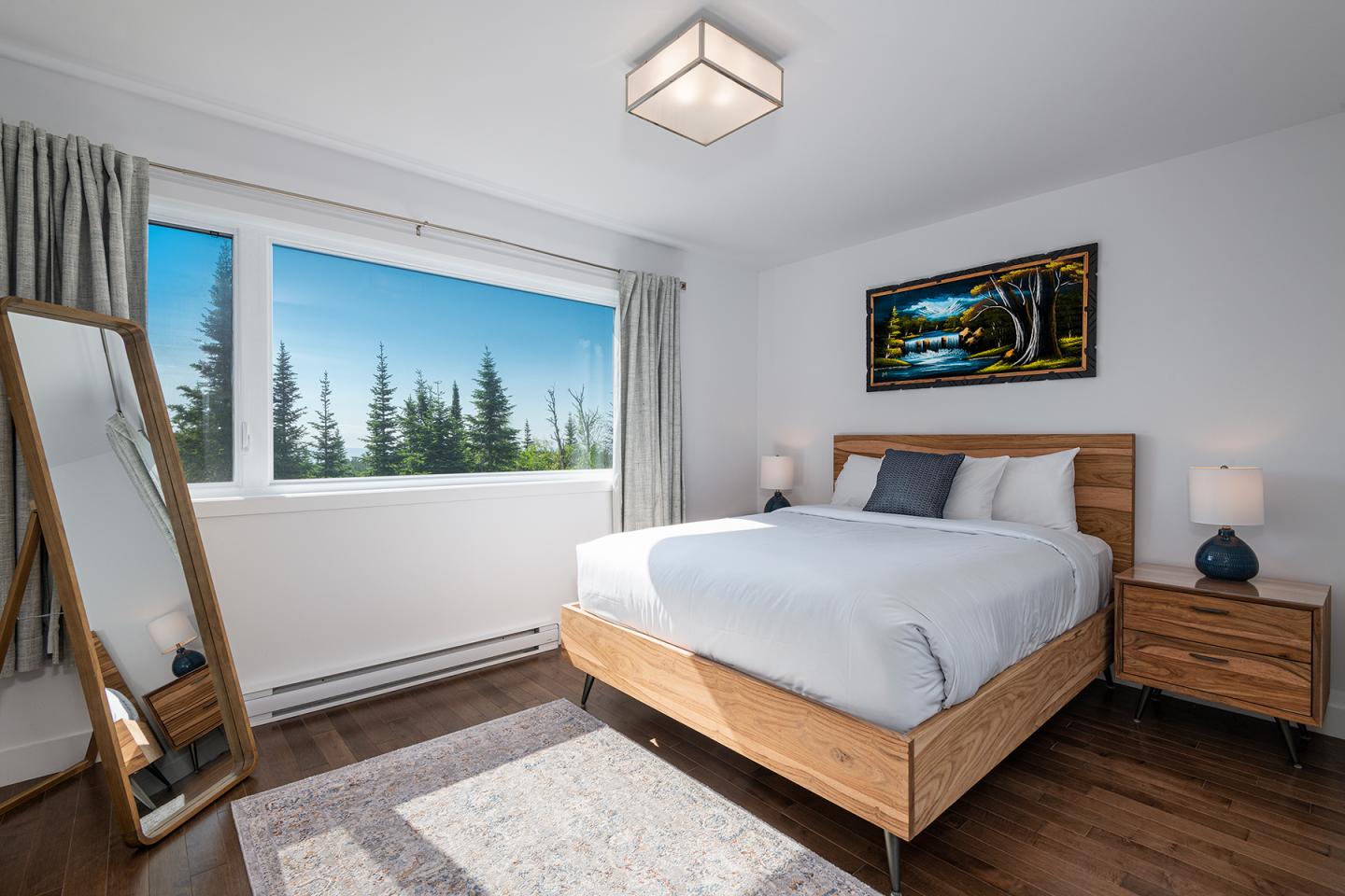 Chambre moderne avec lit double, miroir, grande fenêtre et nature en arrière-plan.