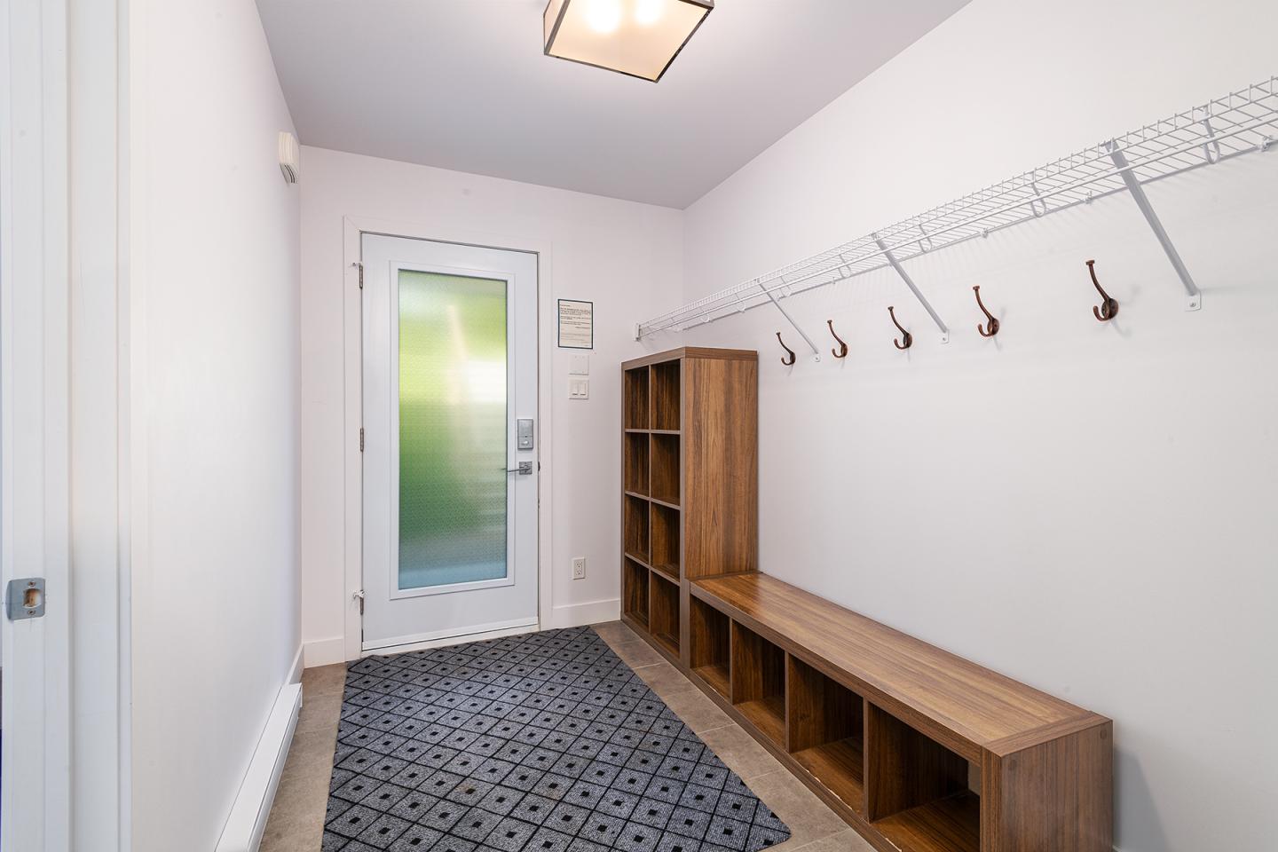 Couloir blanc avec patères, banc en bois et tapis noir.