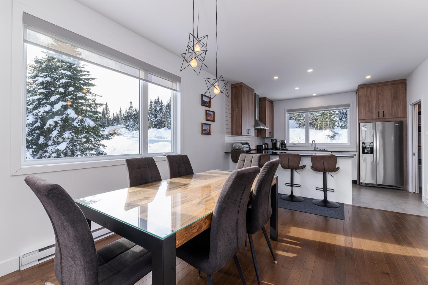 Salle à manger moderne avec table en bois, baies vitrées et cuisine à l'arrière.