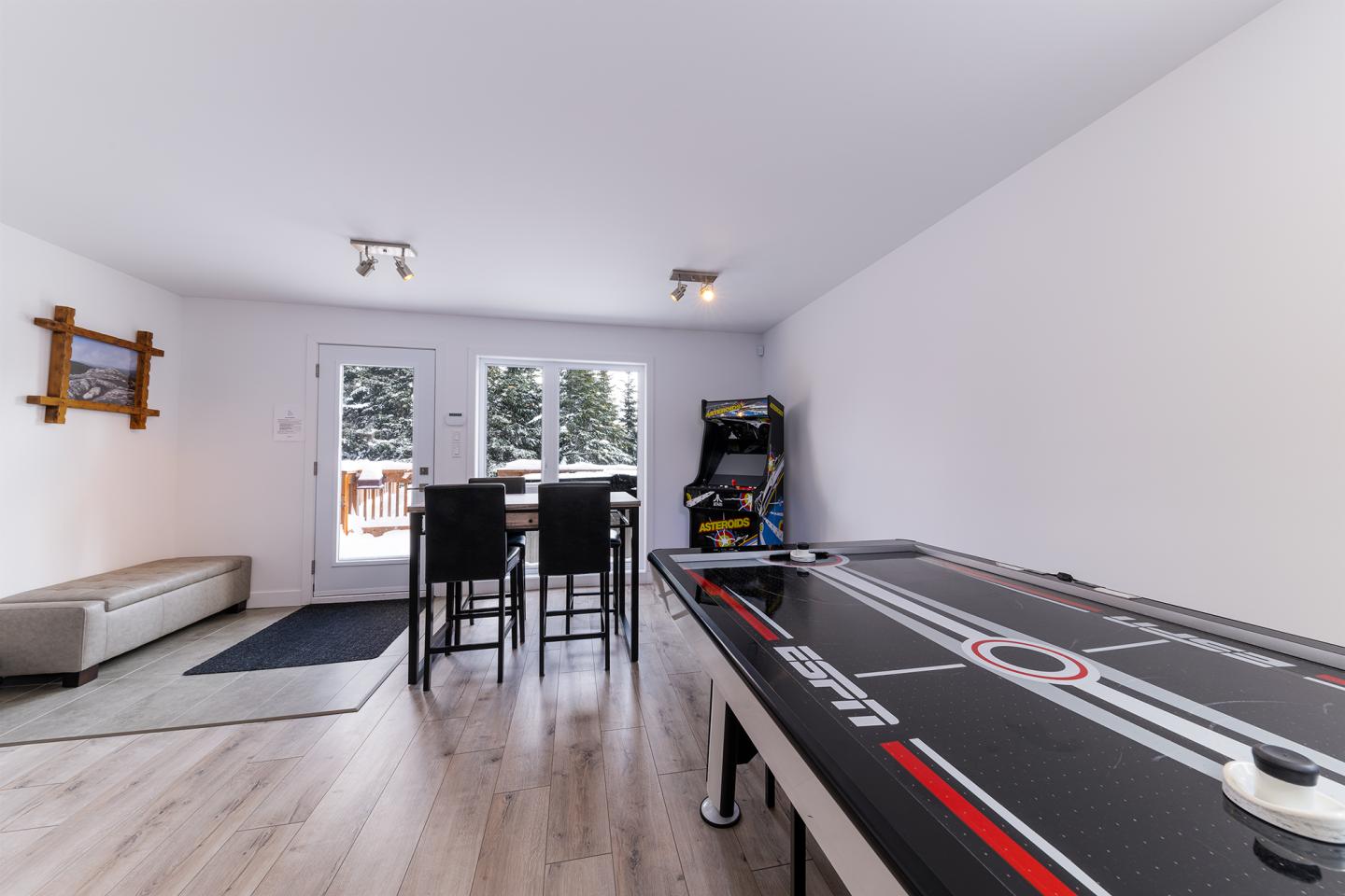 Salle de jeux avec table d'air hockey, arcade, tabourets et vue sur l'extérieur enneigé.