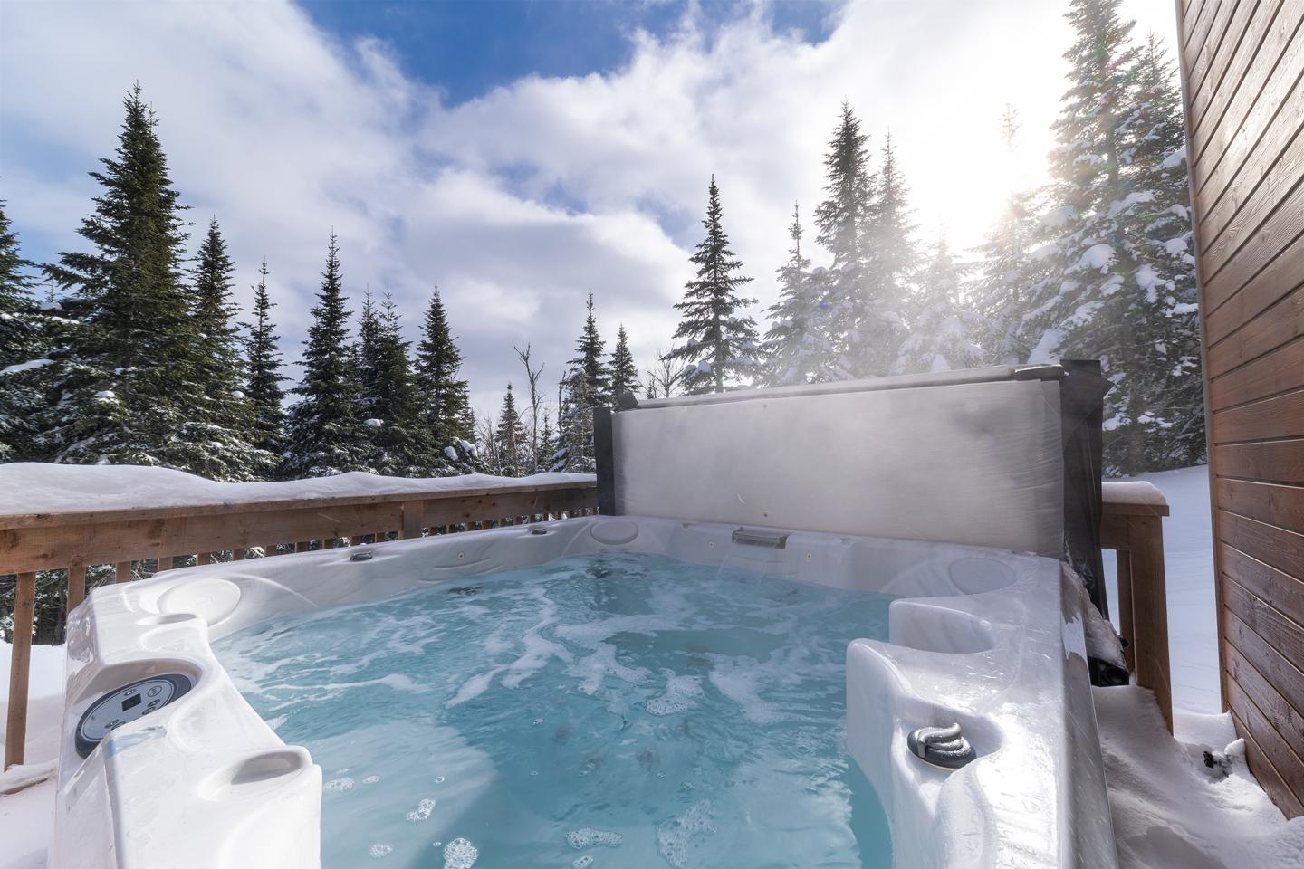 Jacuzzi fumant entouré de neige, ciel bleu et sapins en arrière-plan.