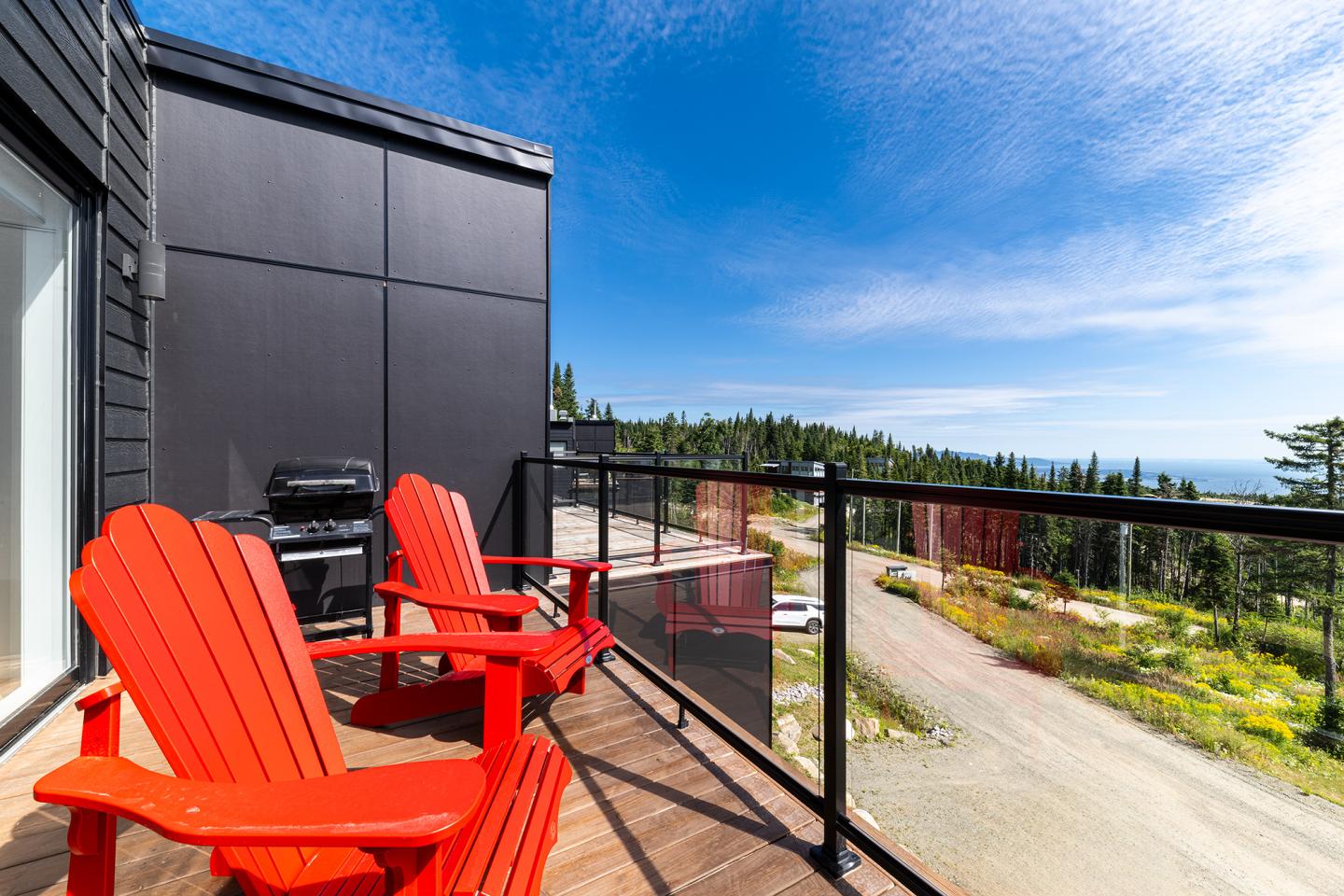 Terrasse avec chaises rouges, barbecue et vue sur forêt sous ciel bleu.
