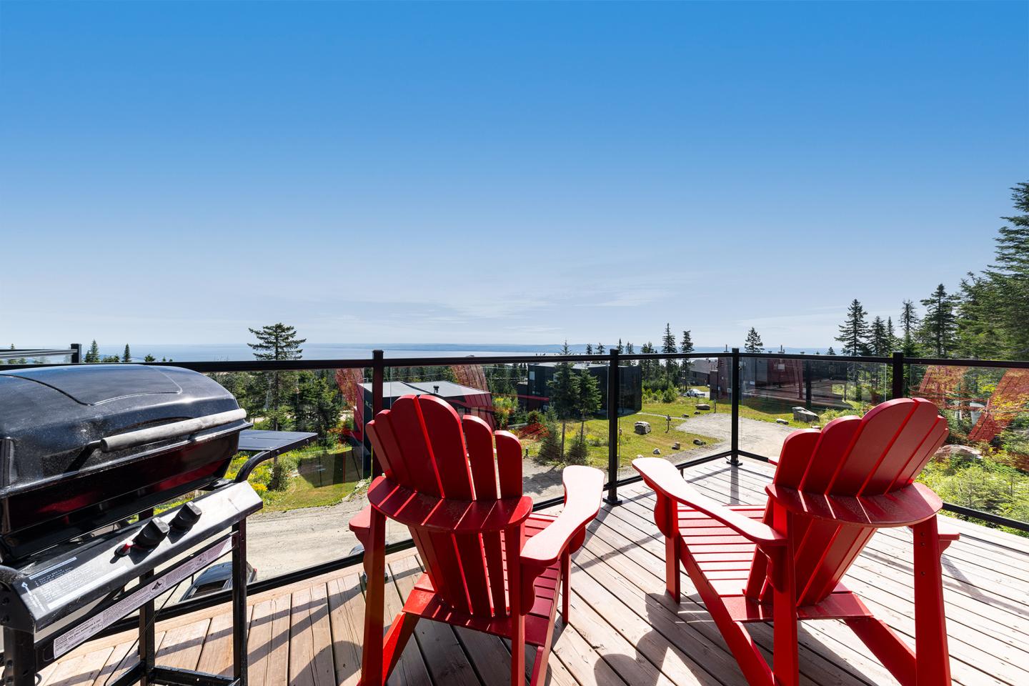 Terrasse ensoleillée avec chaises rouges et barbecue, vue sur jardin et océan.
