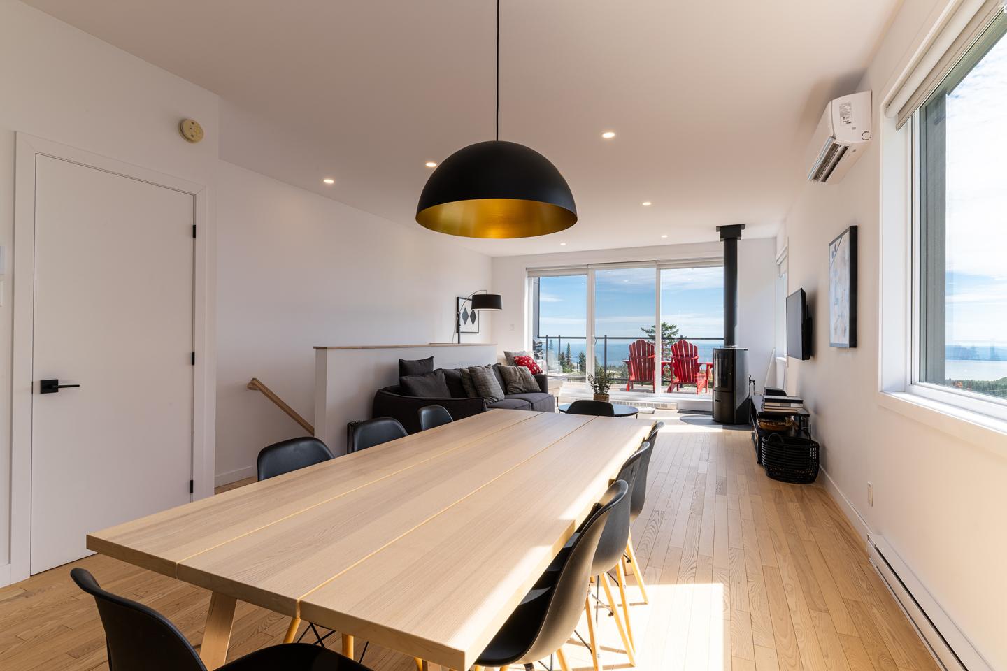 Salle à manger moderne avec table en bois, salon au fond et vue sur la mer.