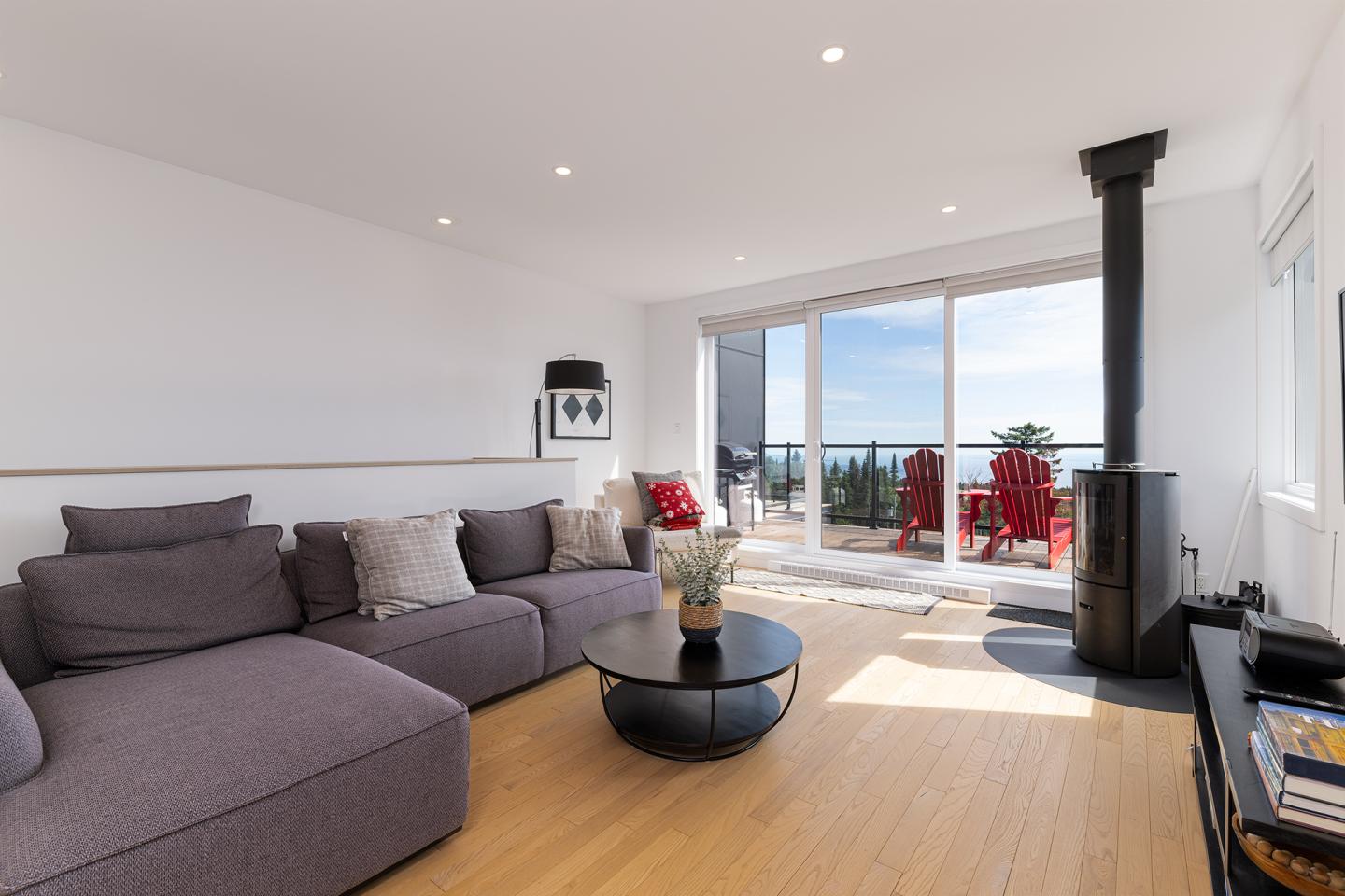 Salon moderne avec canapé gris, poêle noir, baies vitrées et vue sur mer.