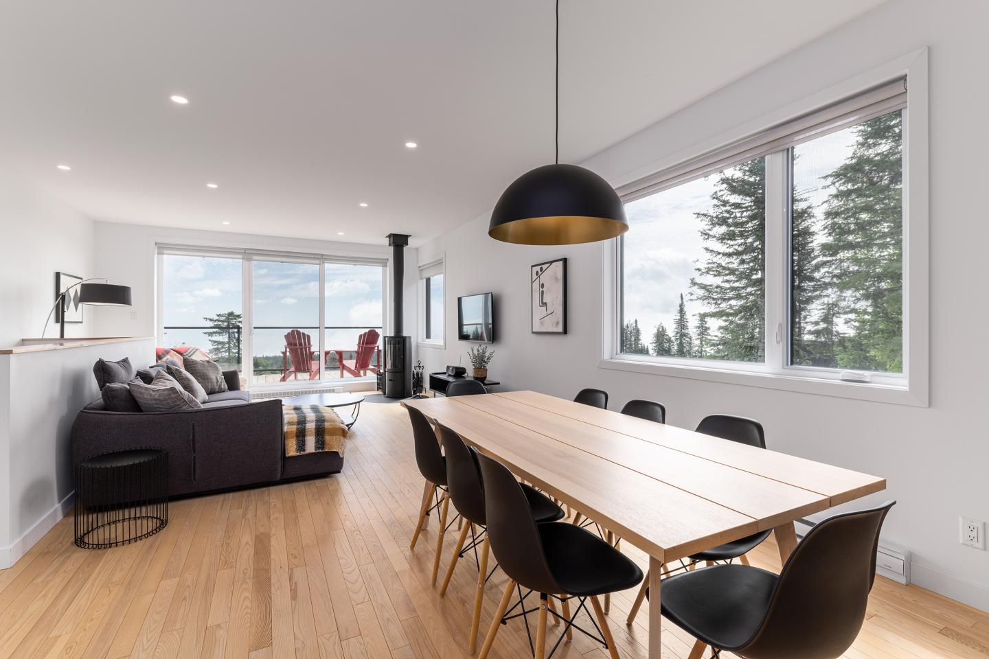 Salle à manger moderne avec table en bois et lampes noires, vue sur salon et baies vitrées.
