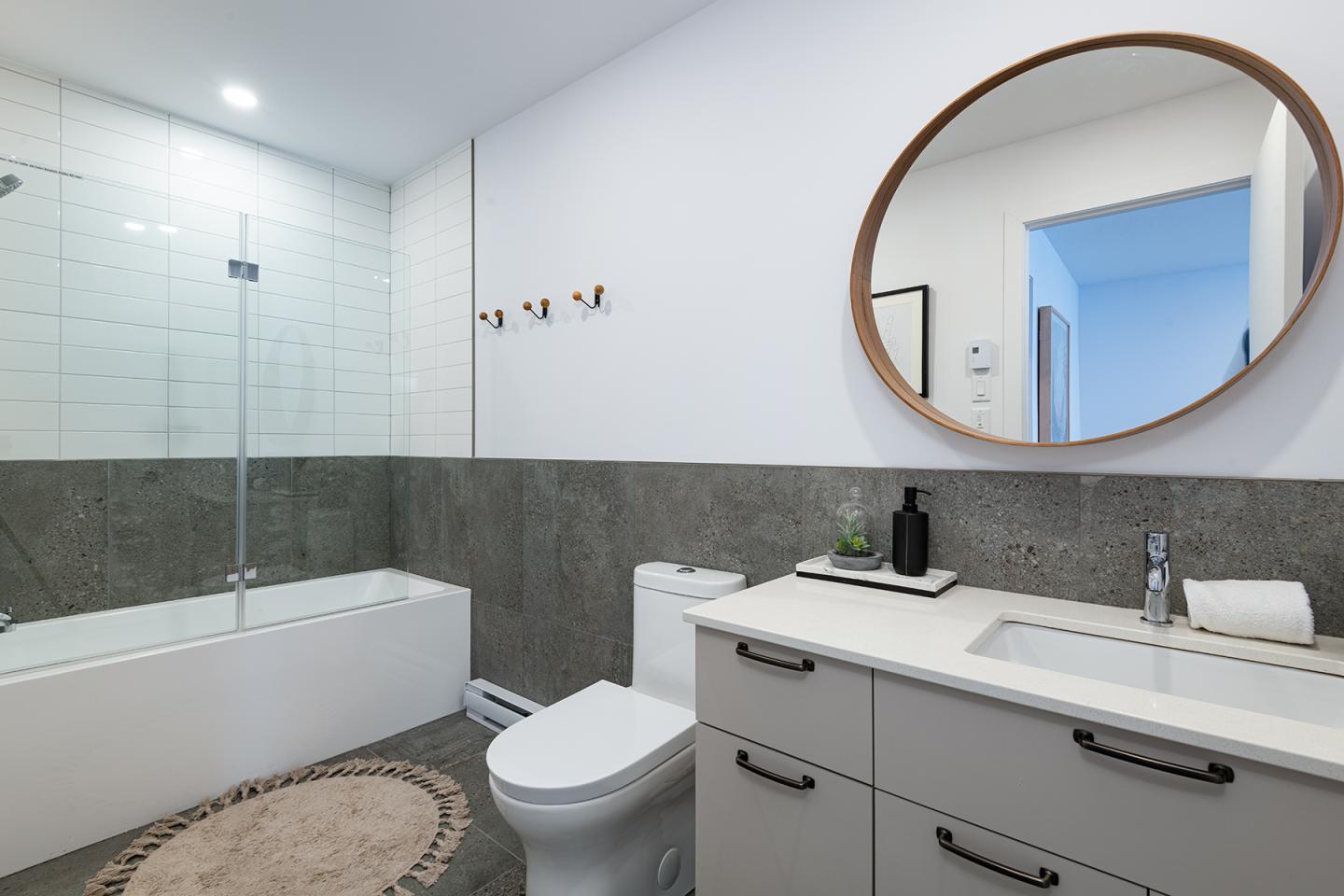 Salle de bain moderne avec baignoire, lavabo, miroir rond et carrelage gris.