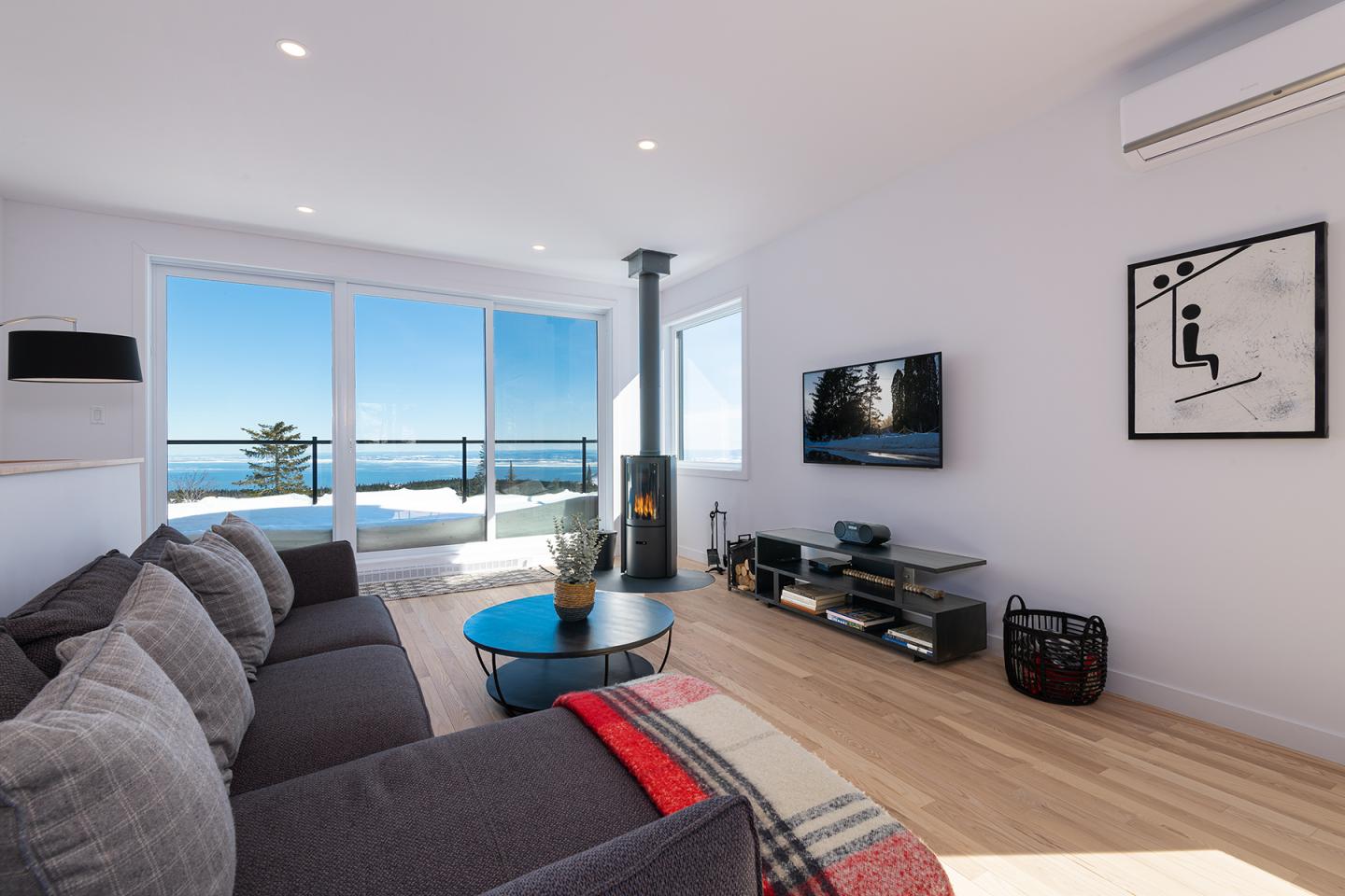 Salon moderne avec vue sur mer, canapé gris, et télévision.