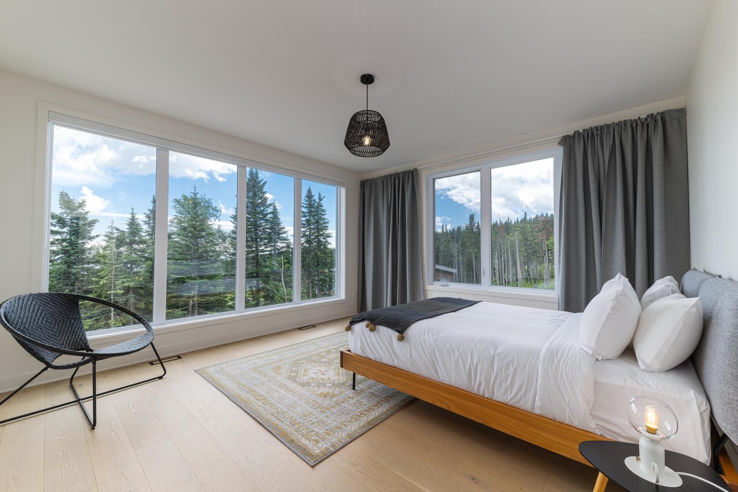 Chambre lumineuse avec lit, fauteuil et grandes fenêtres donnant sur une forêt.