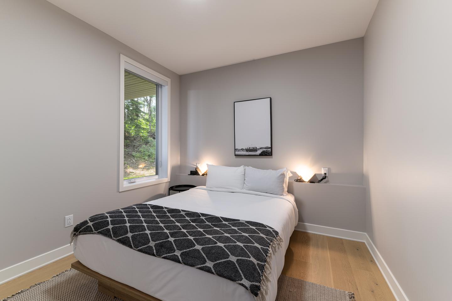 Chambre minimaliste avec un lit double, murs gris, et fenêtre avec vue sur la verdure.