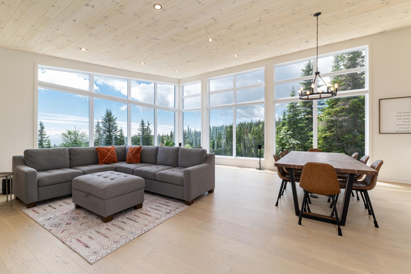 Salon lumineux avec canapé gris, grande table à manger, fenêtres sur forêt, intérieur moderne.
